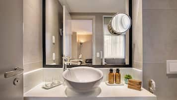 A modern bathroom with a large bathtub, a round mirror, and a vanity with a bowl sink. There are toiletries on the vanity and a potted plant beside the sink.