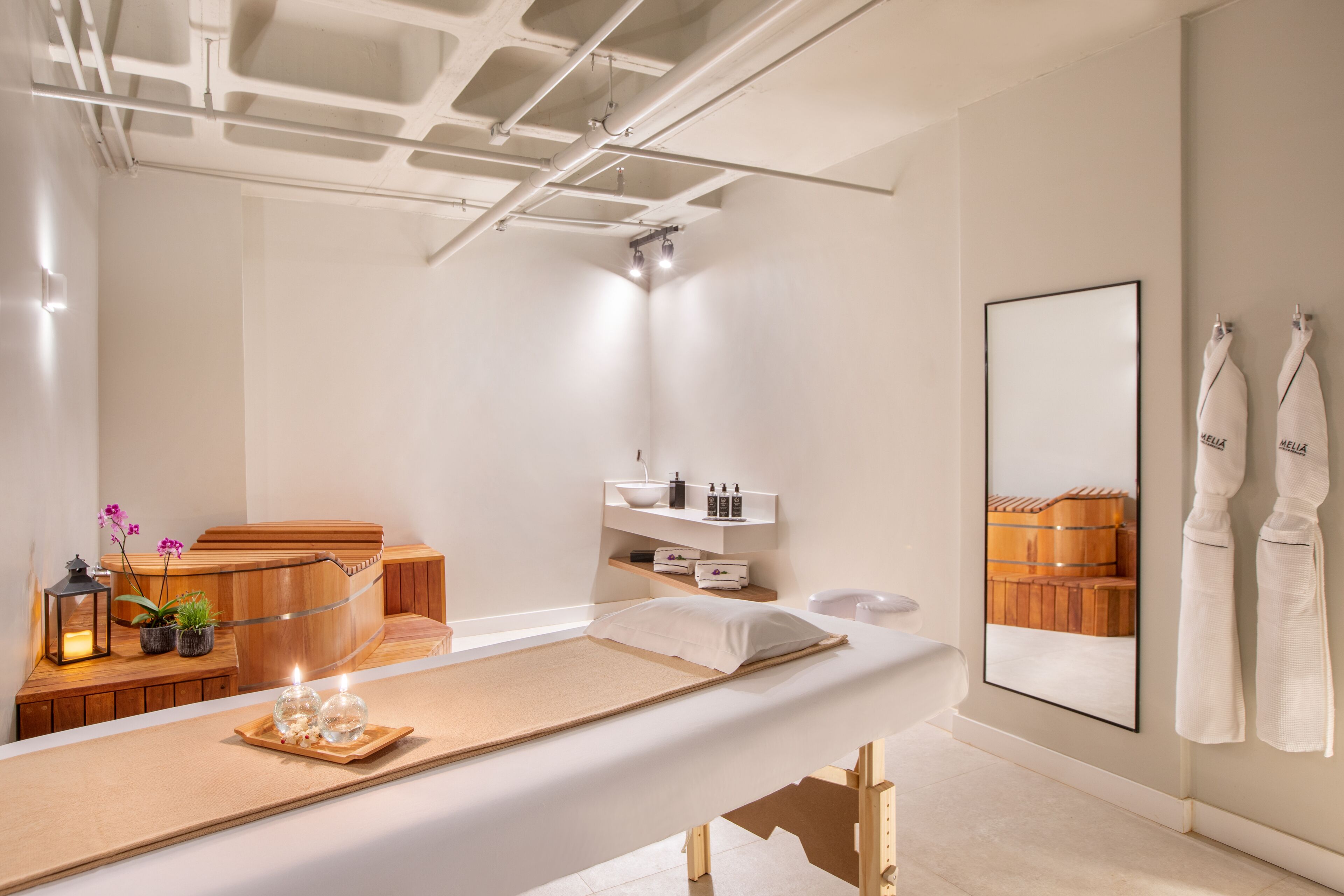 A spa room with a massage table, wooden bench, shelves with toiletries, and a wooden sauna visible in the reflection.