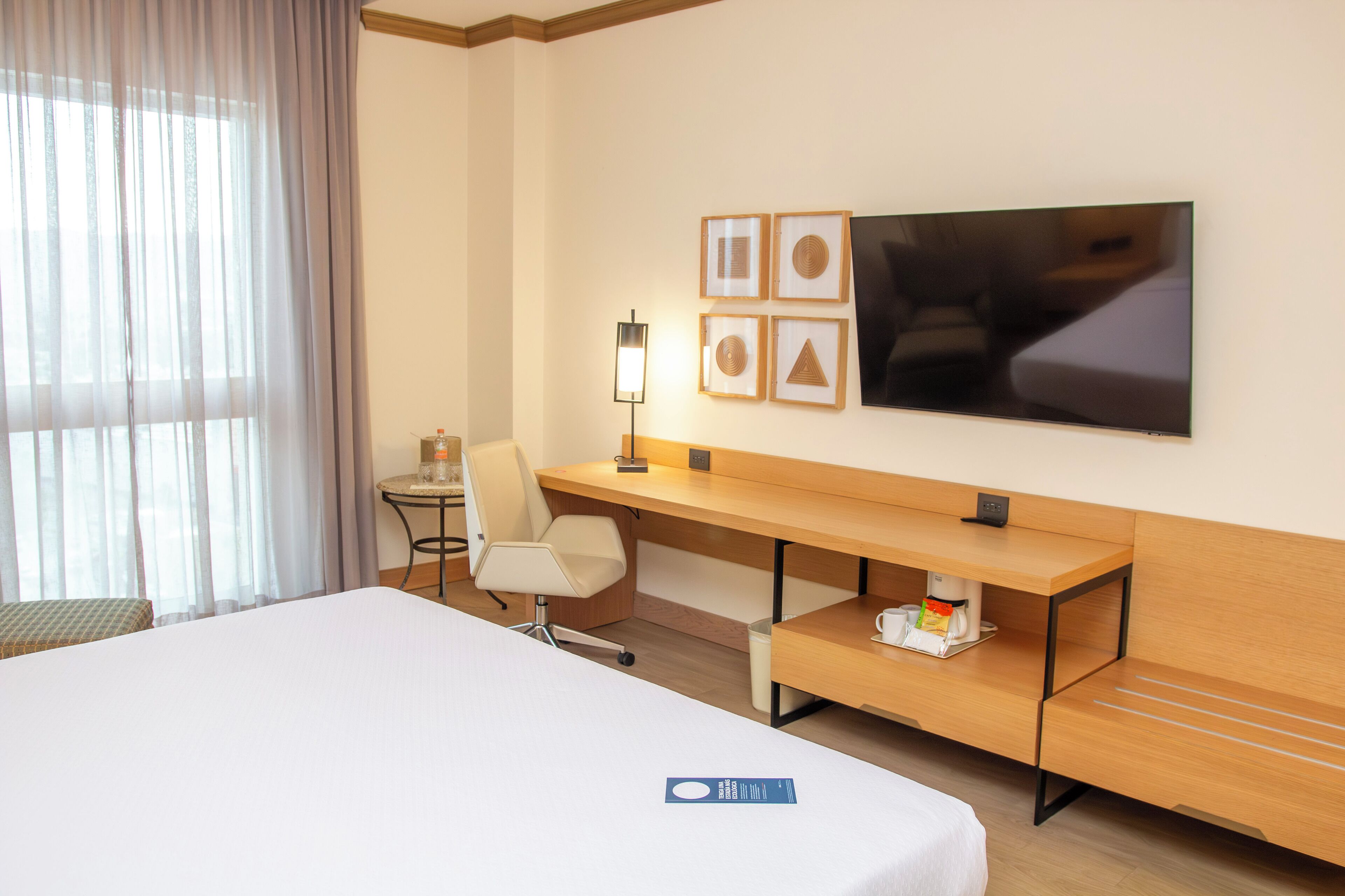 A hotel room with a bed, a desk, a chair, a TV, and a small table with a lamp.