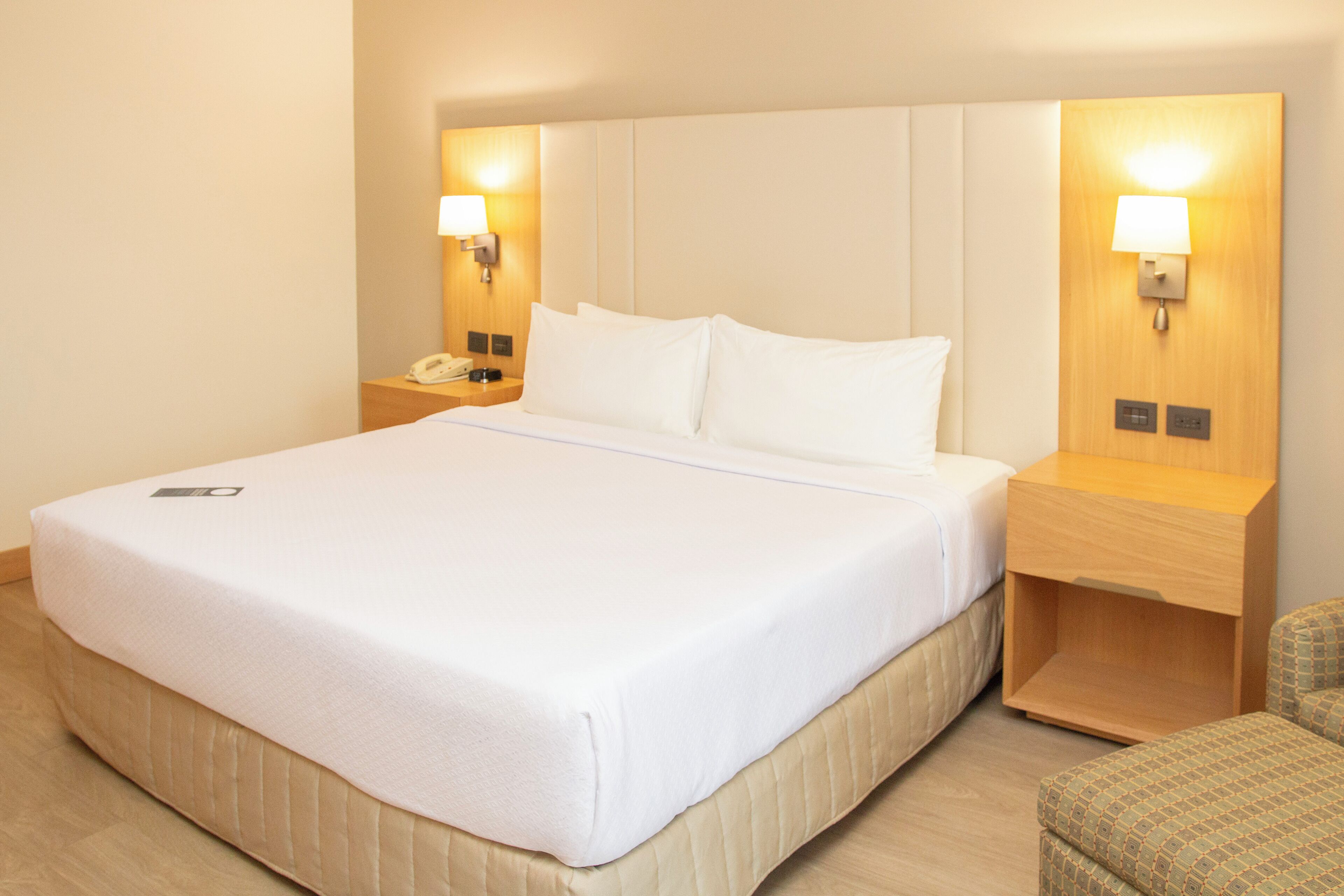 Una habitación de hotel con una cama grande, lámparas de noche, un teléfono en la mesita y una pequeña silla.