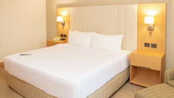 Una habitación de hotel con una cama grande, lámparas de noche, un teléfono en la mesita y una pequeña silla.