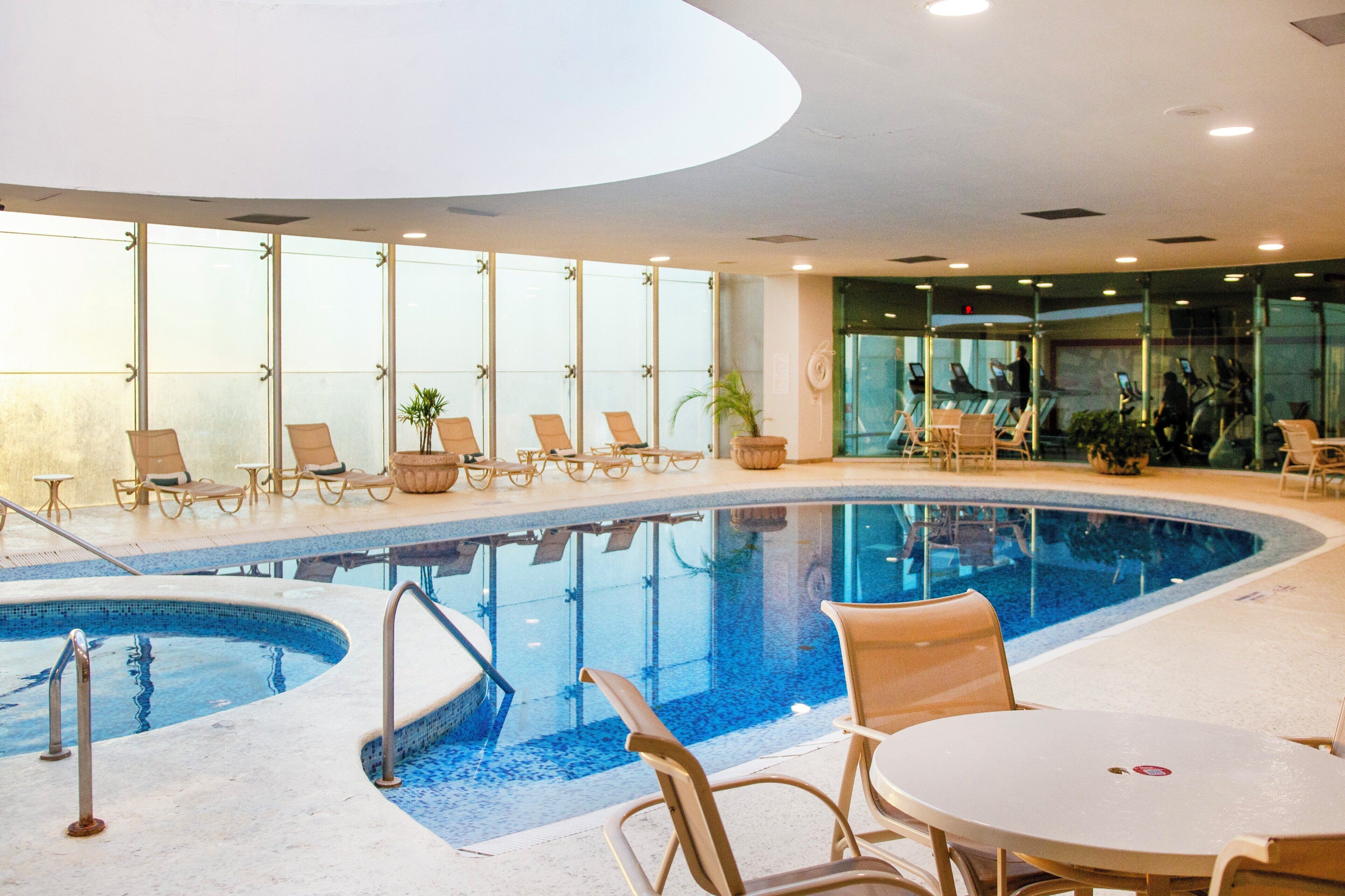 An indoor swimming pool with lounge chairs and a table, surrounded by large windows.
