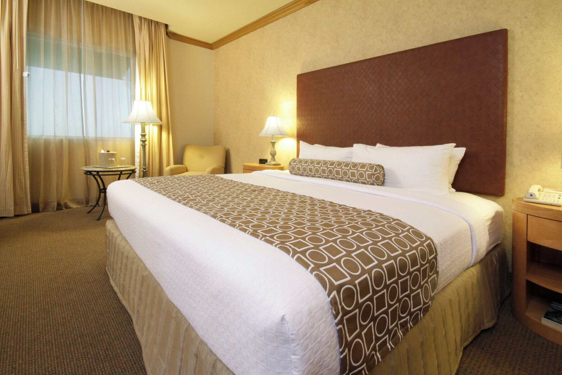 A hotel room with a large bed, two bedside lamps, a chair, a small table, and a telephone on a wooden nightstand.