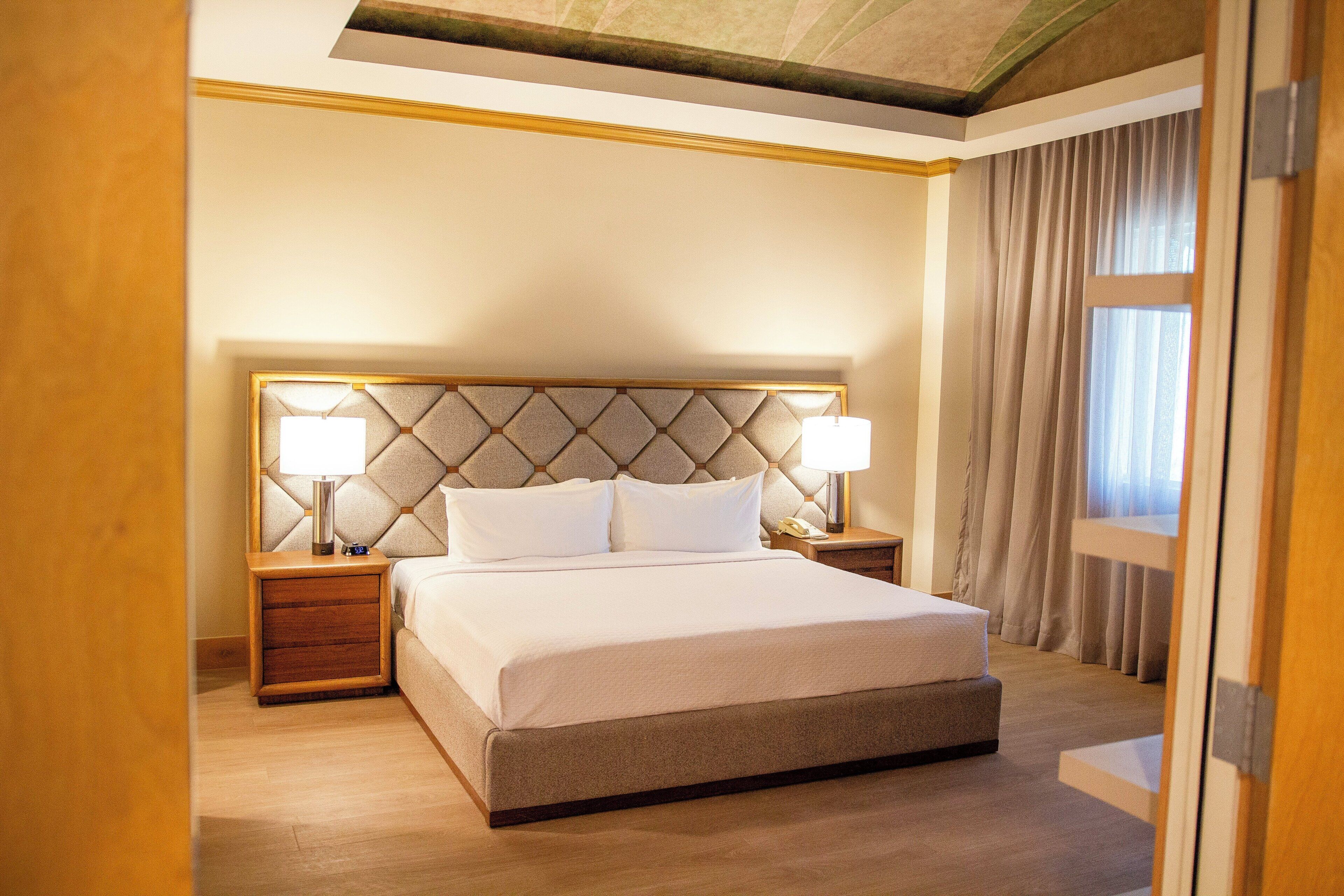 A hotel room with a large bed, two bedside tables, a headboard, and a window with curtains.
