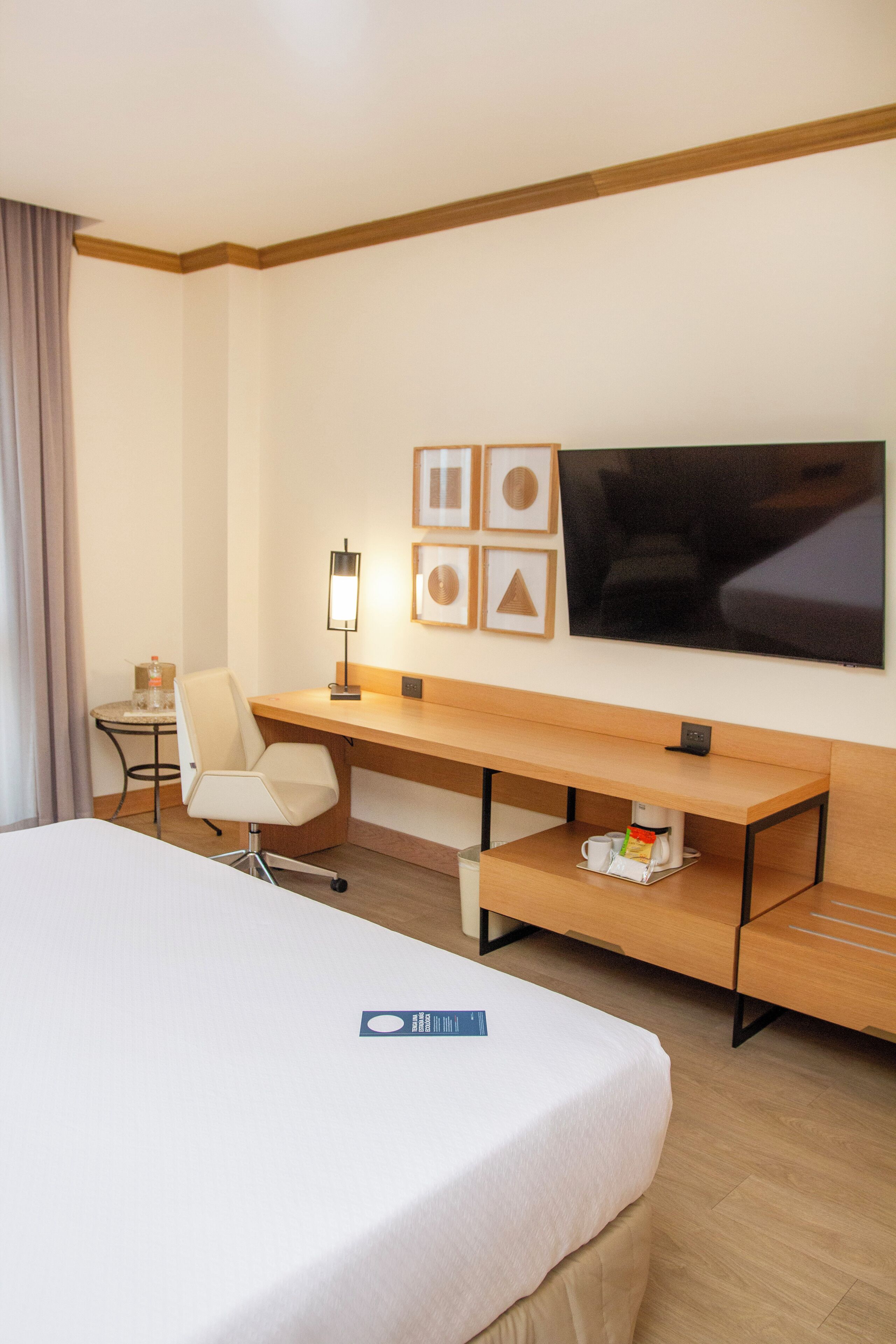 A hotel room with a bed, a desk, a chair, a TV, and a small table with a lamp and a bottle.