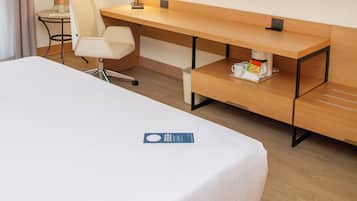 A hotel room with a bed, a desk, a chair, a TV, and a small table with a lamp and a bottle.