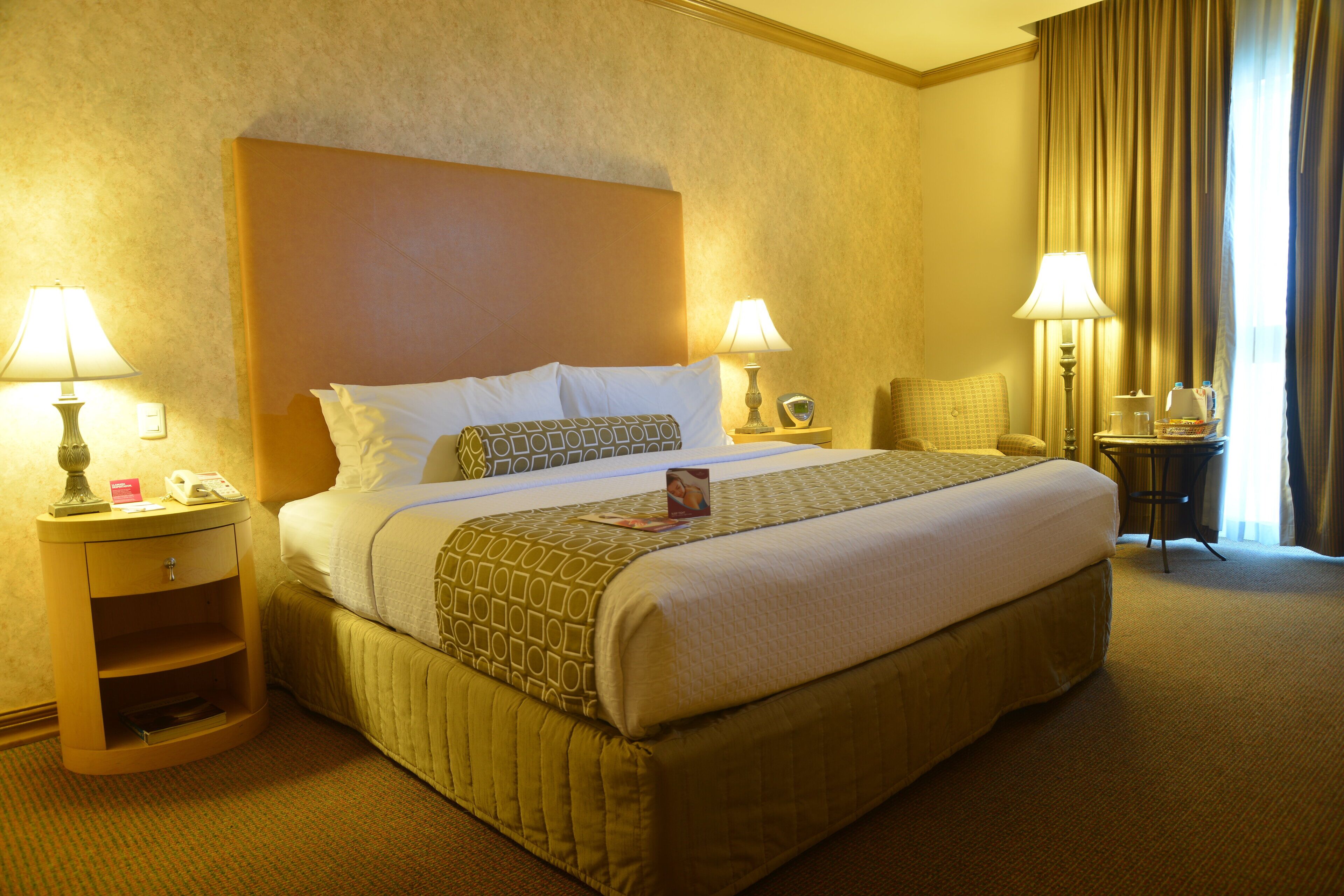 Una habitación de hotel con una cama grande, dos mesitas de noche con lámparas, una silla y una pequeña mesa con lámpara y una taza.