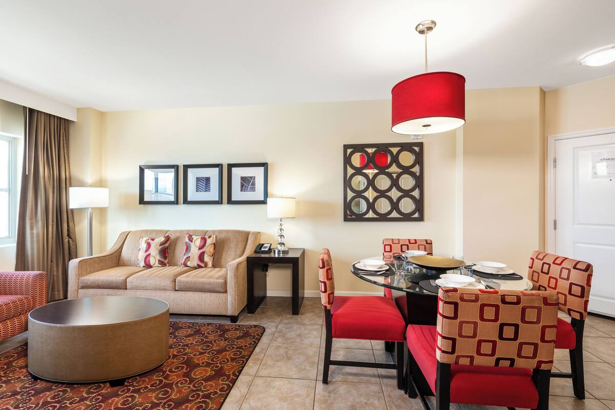 A modern living room with a sofa, a round coffee table, and a dining area with red chairs and a glass table.