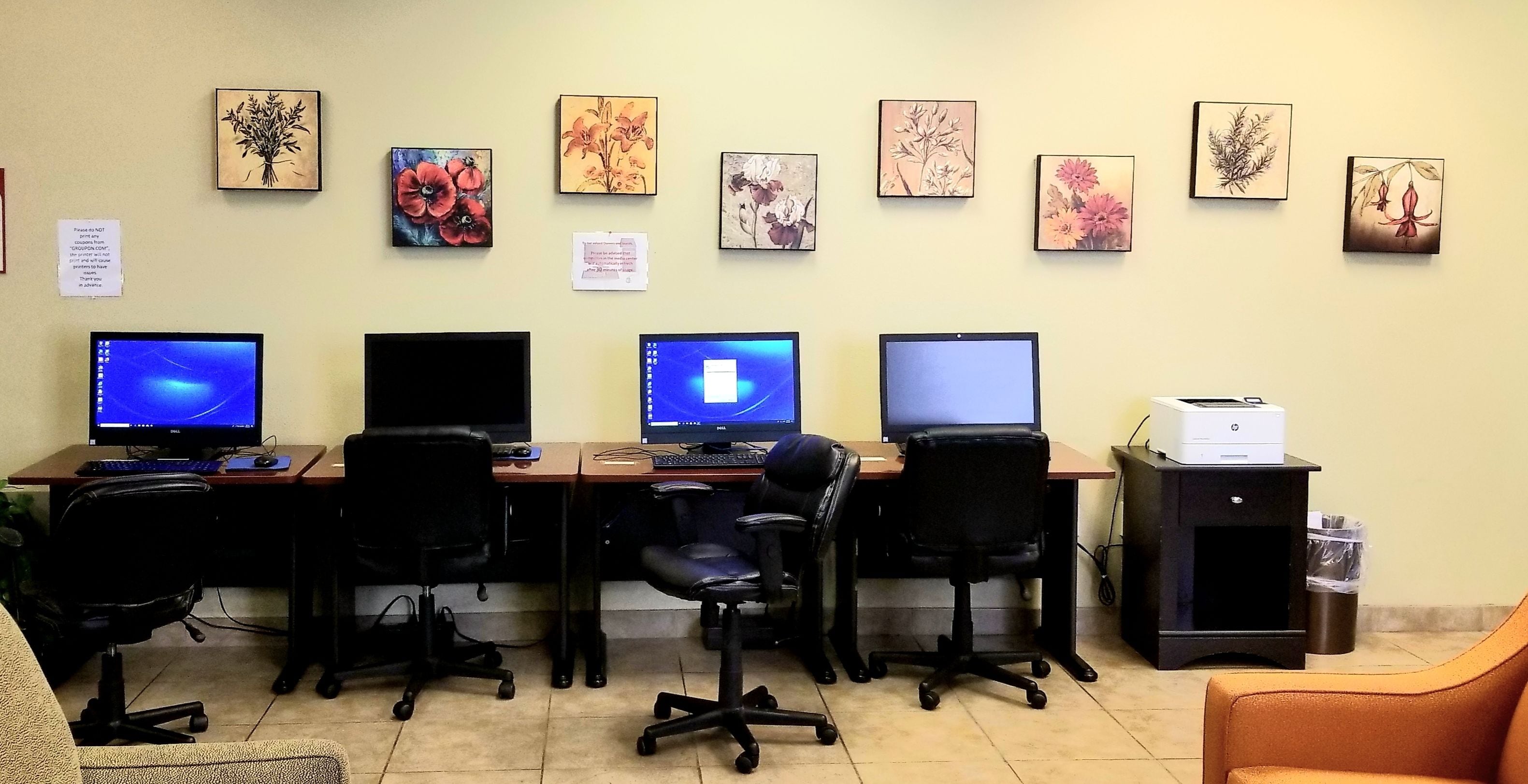 A workspace with four desks, each with a computer and a chair. There are floral artworks on the wall and a printer on the right.