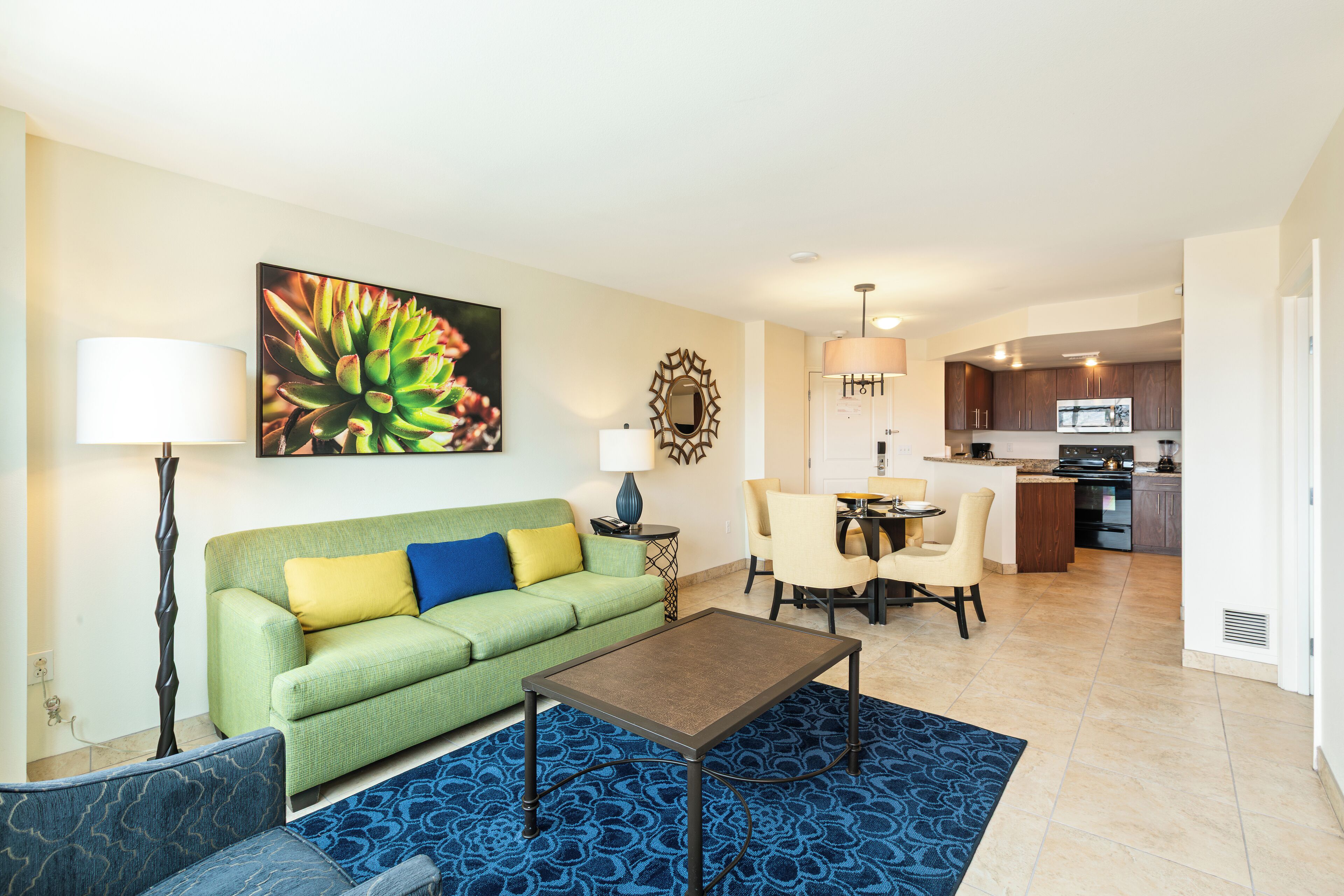 A living room with a green sofa, a coffee table, and a blue patterned rug. There is a dining area with a table and chairs, and a kitchen in the background.