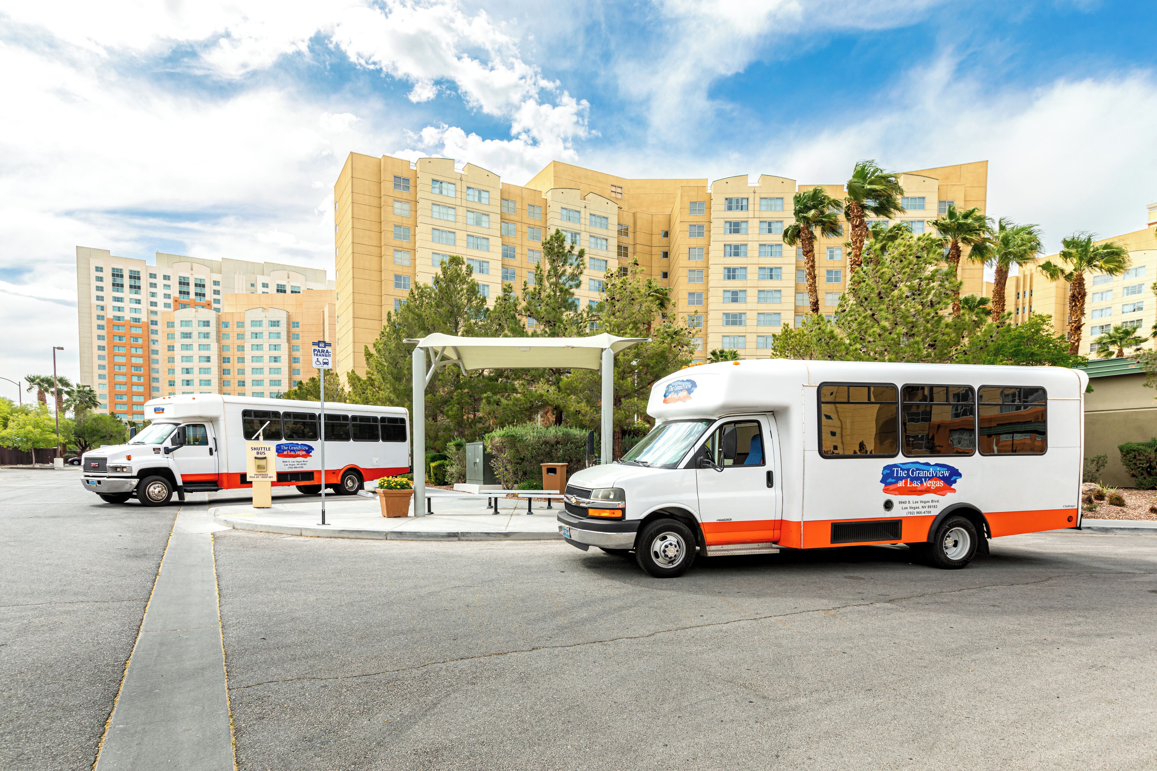 Two white and orange buses parked in front of a hotel with multiple floors and palm trees.