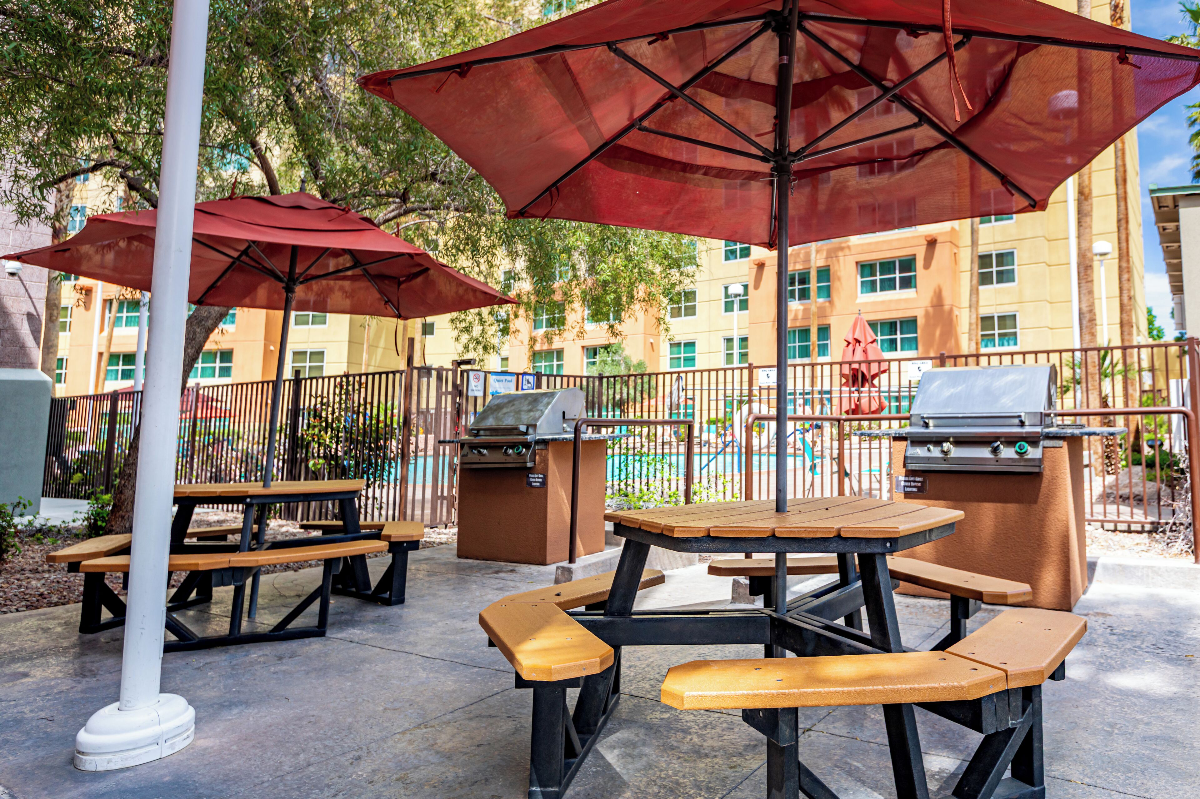 An outdoor patio area with picnic tables and barbecue grills, surrounded by a fence and trees.