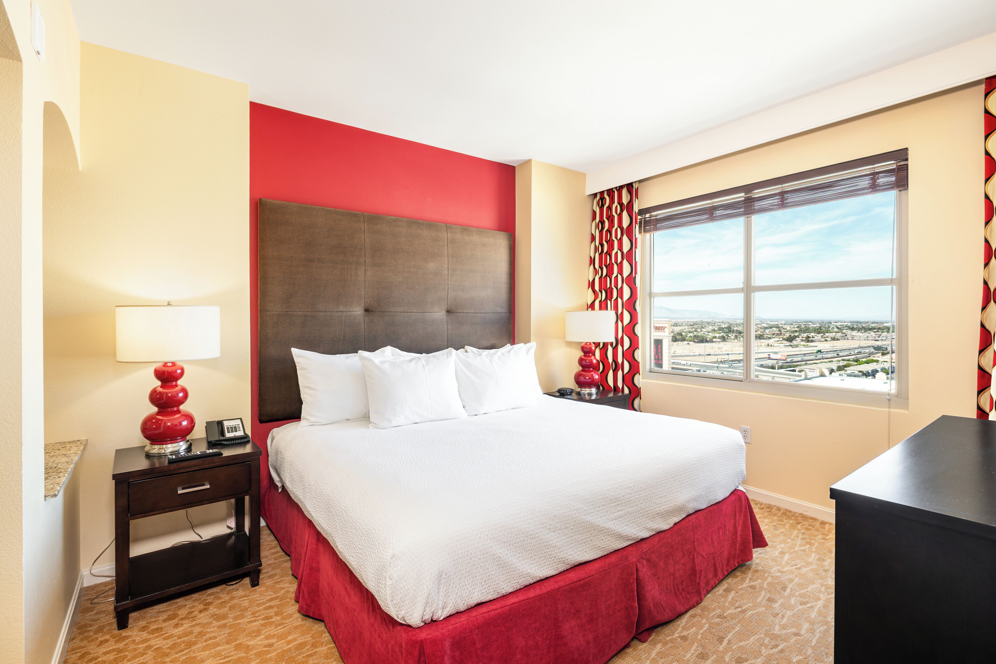 Hotelkamer met een groot bed, uitzicht op de stad, een rode accentmuur en een gordijn met een rood-wit patroon.