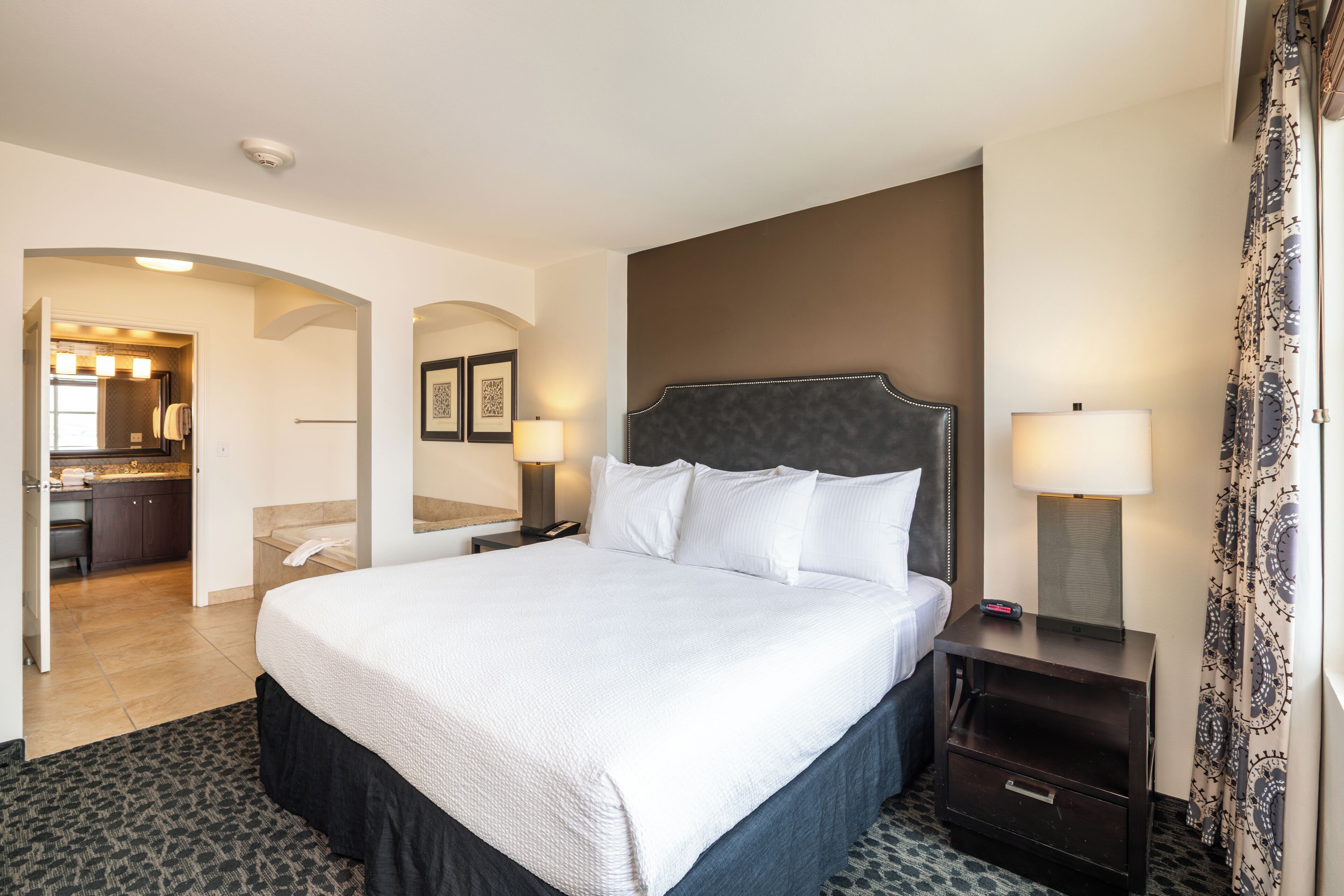 A hotel room with a large bed, a nightstand with a lamp, a desk with a chair, and a bathroom visible through an archway.
