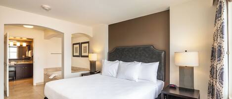 A hotel room with a large bed, a nightstand with a lamp, a desk with a chair, and a bathroom visible through an archway.