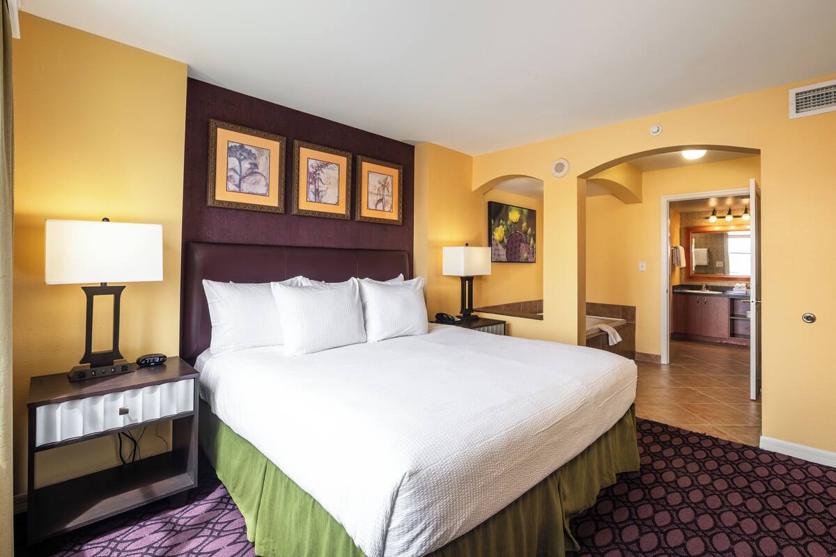 A hotel room with a large bed, two bedside tables, a nightstand, a green bed skirt, and a patterned carpet.