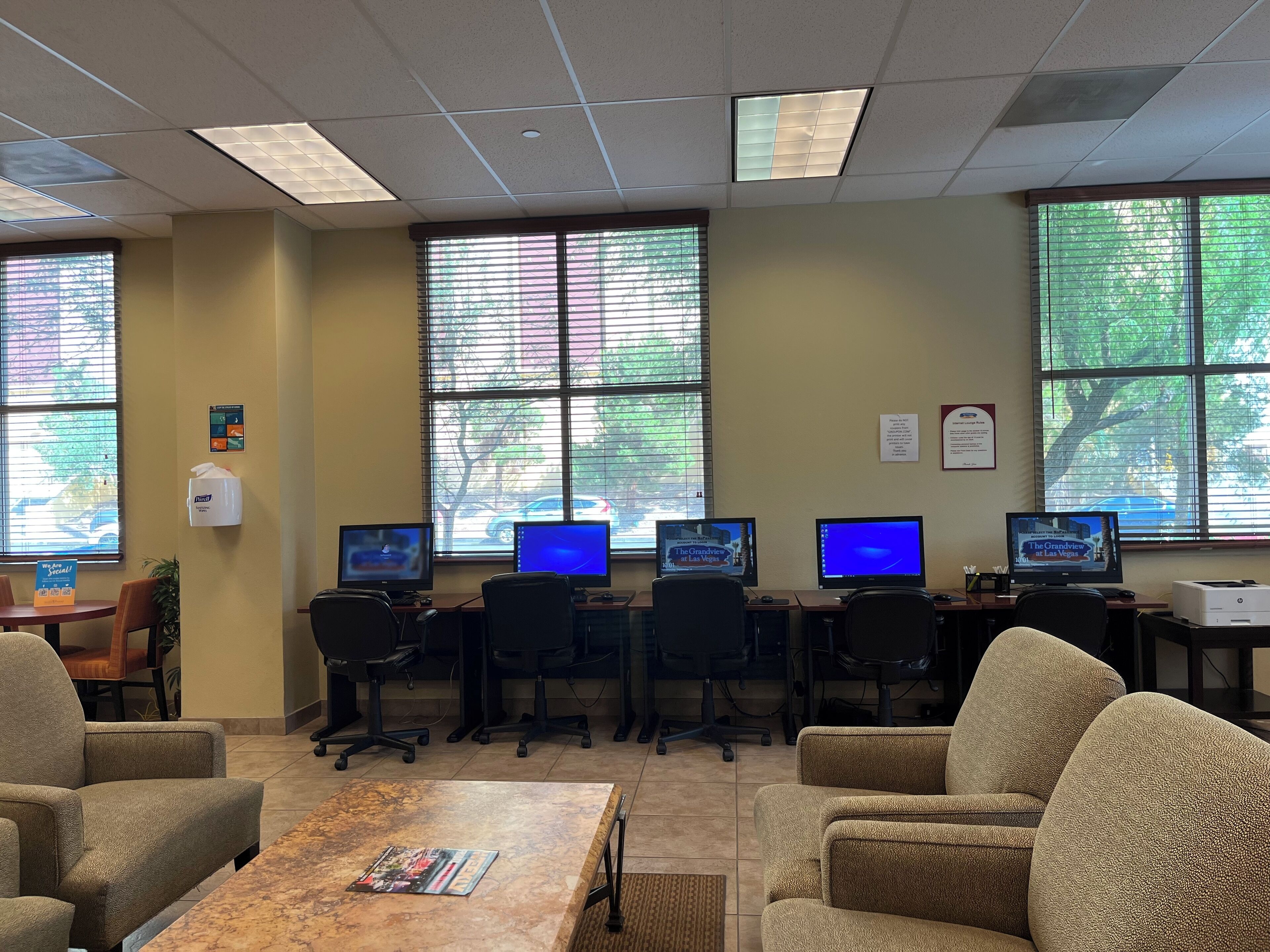 A conference room with multiple computer monitors, office chairs, a coffee table, and large windows with blinds.