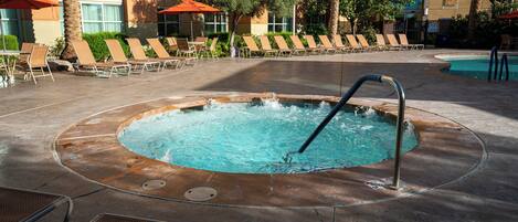 A hot tub surrounded by lounge chairs and umbrellas, with a pool in the background.
