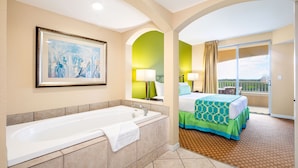 Habitación de hotel con bañera, cama con colcha verde y azul, balcón con vistas y un cuadro en la pared.