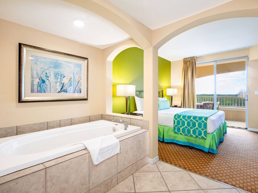 Habitación de hotel con bañera, cama con colcha verde y azul, balcón con vistas y un cuadro en la pared.