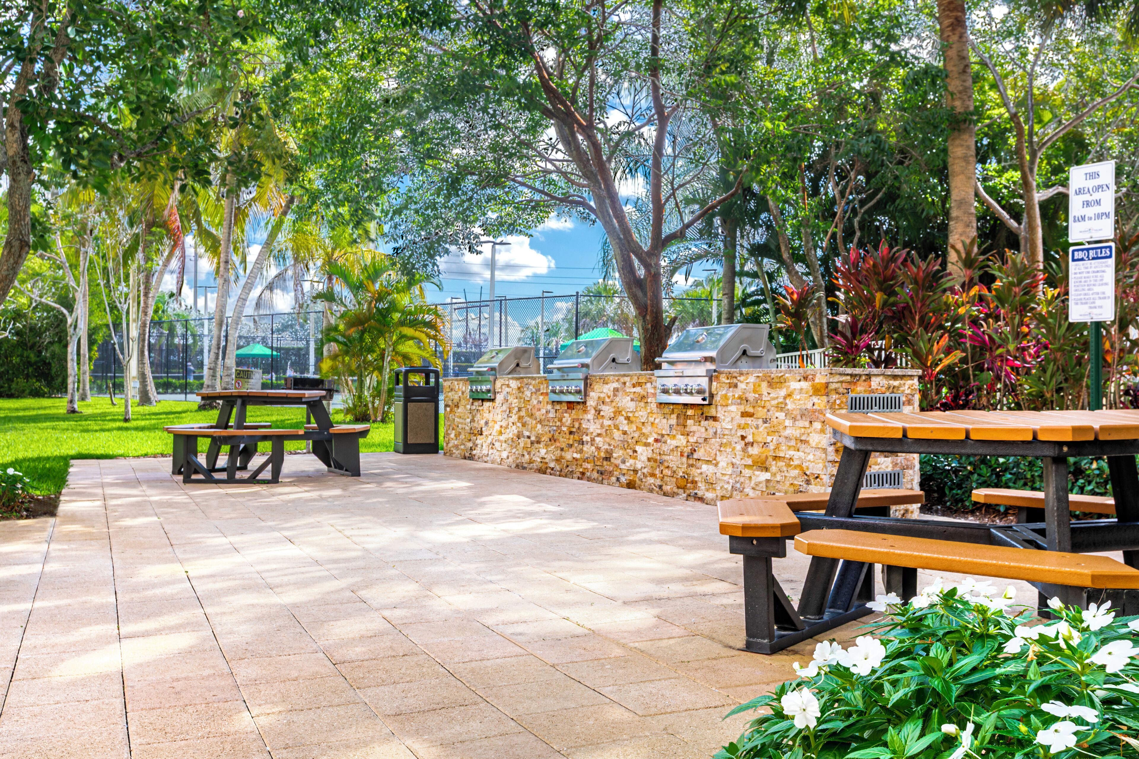 Un espace extérieur aménagé en terrasse, avec des tables de pique-nique et des barbecues, entouré d’une végétation luxuriante et d’un mur en pierre.