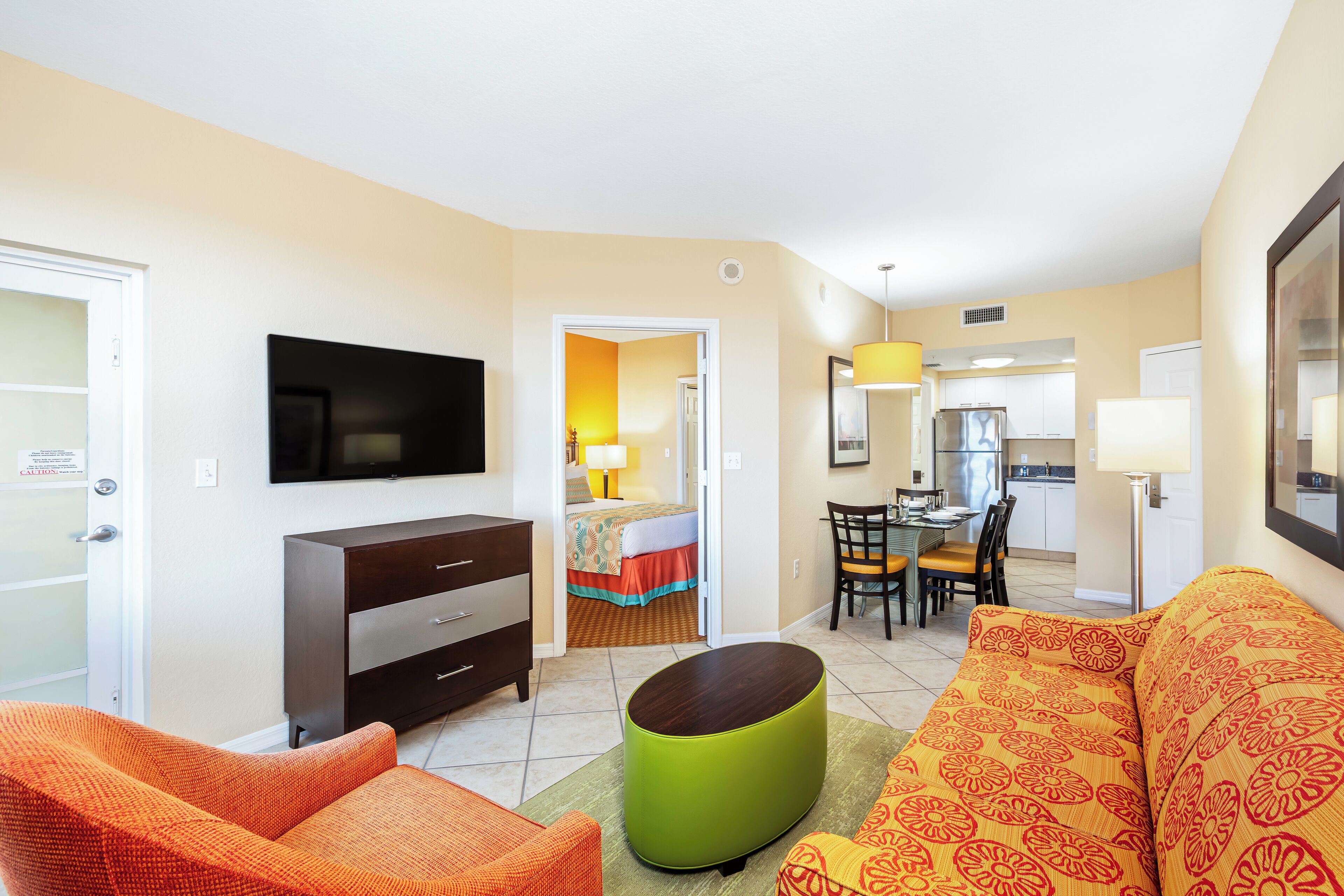 Habitación de hotel con televisor, sofá, mesa de comedor con sillas, zona de cocina y cama con colcha colorida.