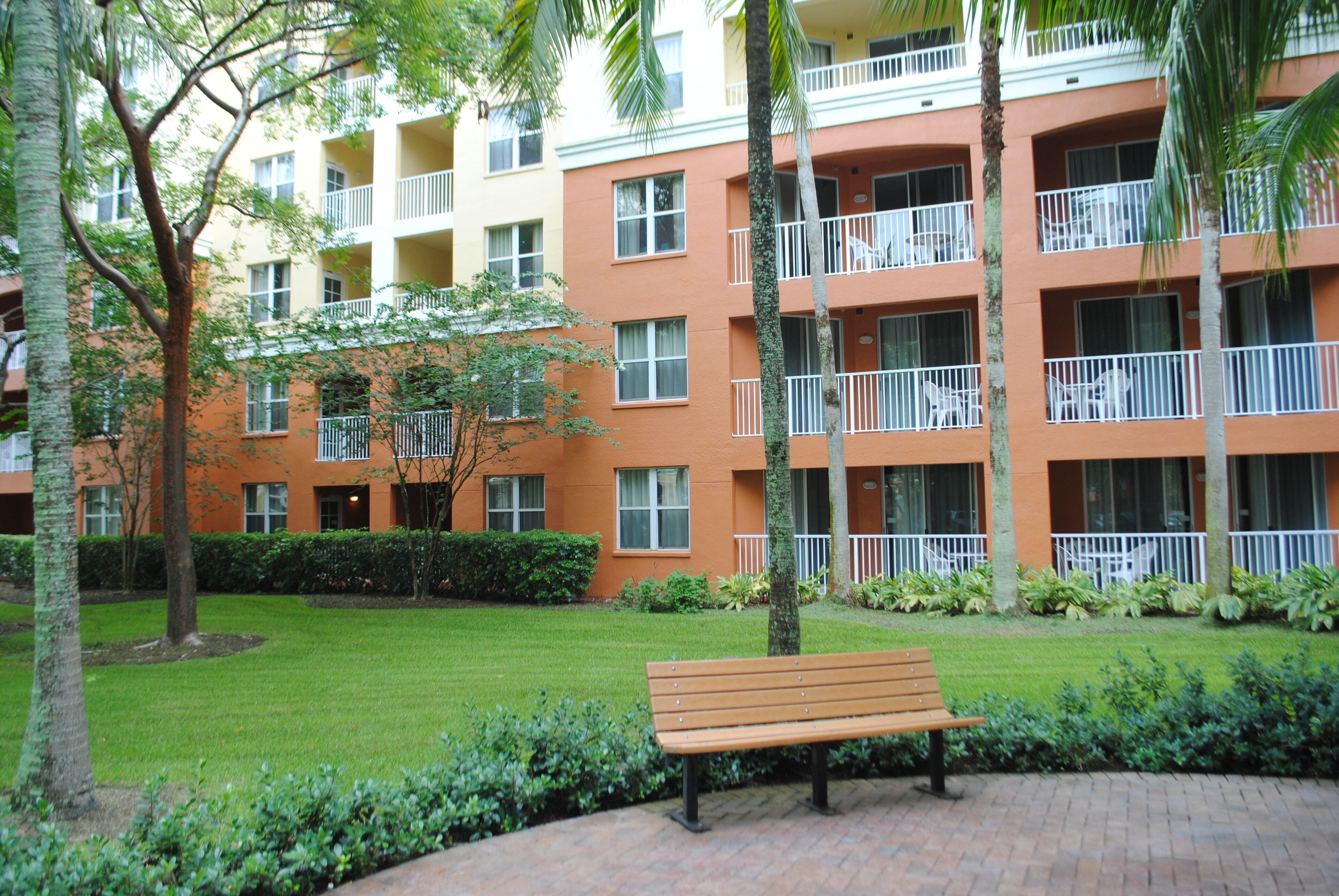 A multi-story building with balconies, a grassy courtyard, and a wooden bench.