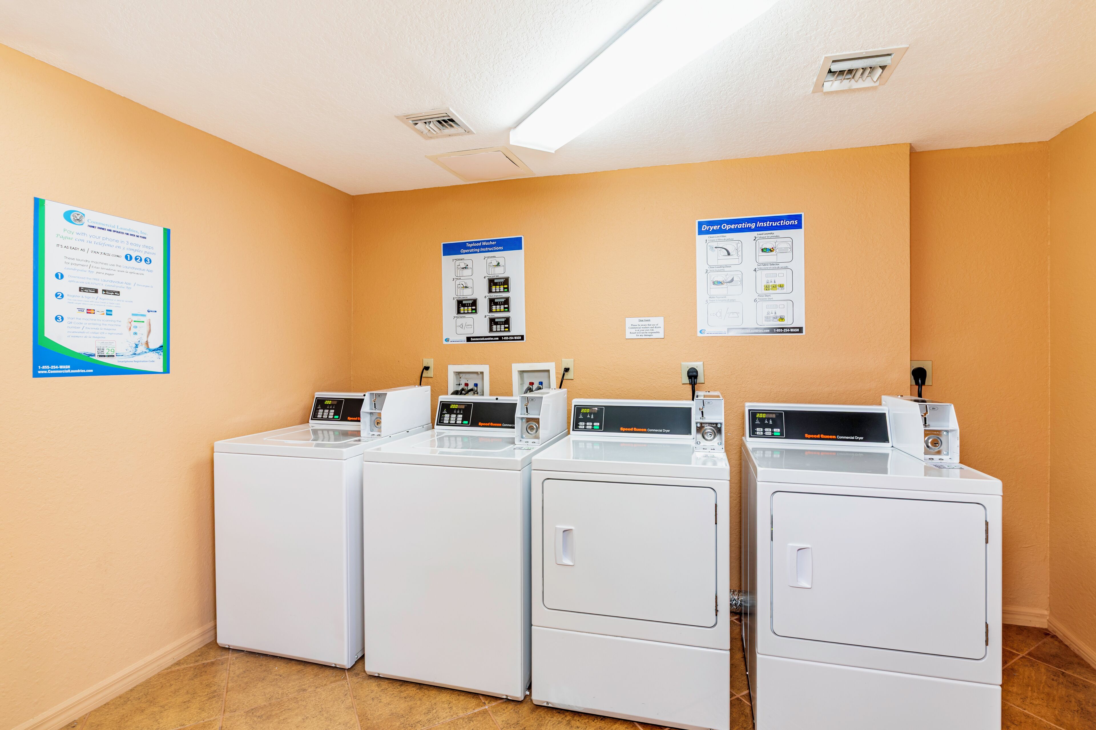 A laundry facility with four white commercial dryers, operating instructions on the wall, and a poster with payment information.