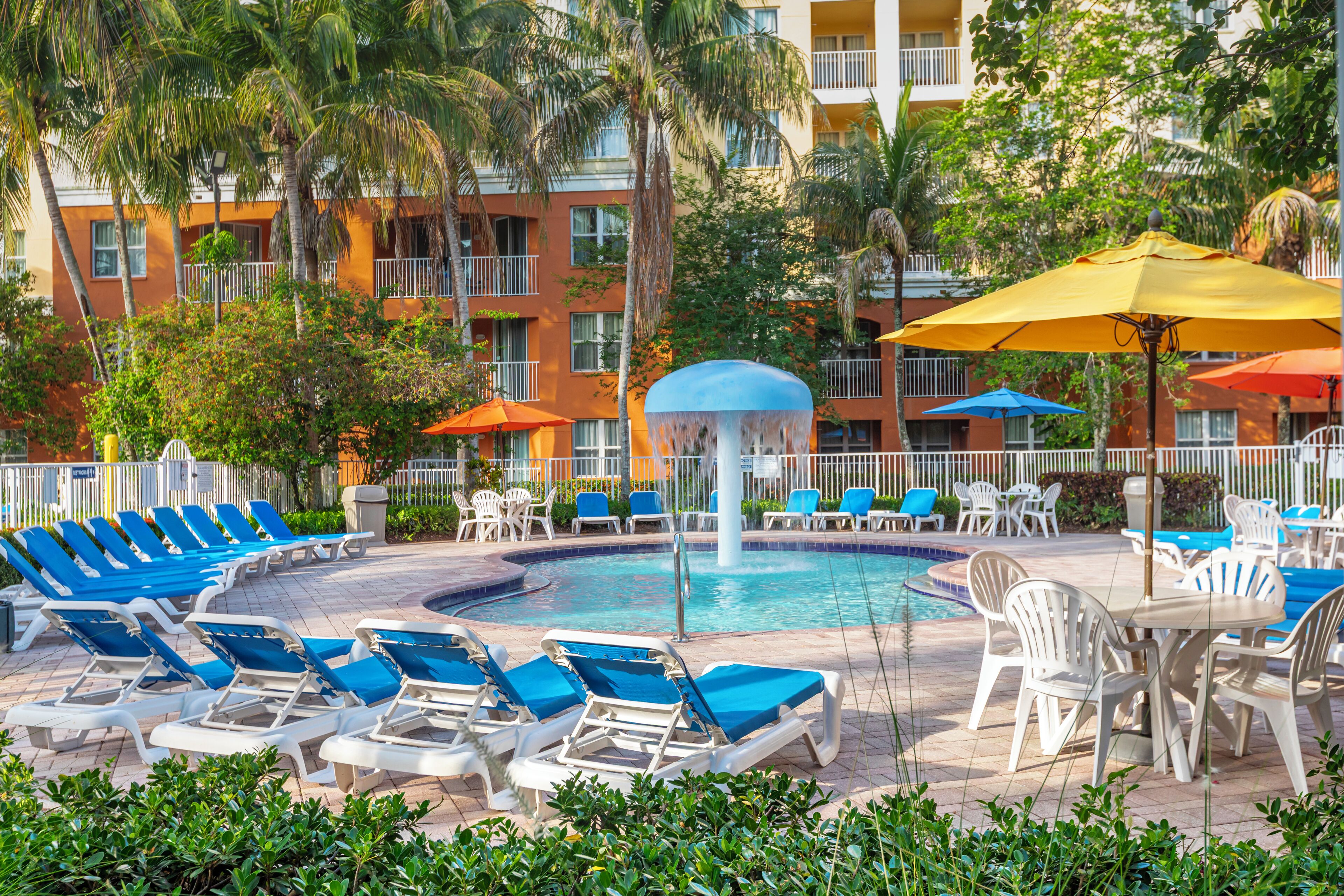 A poolside area with lounge chairs and umbrellas, surrounded by a building with balconies and palm trees.