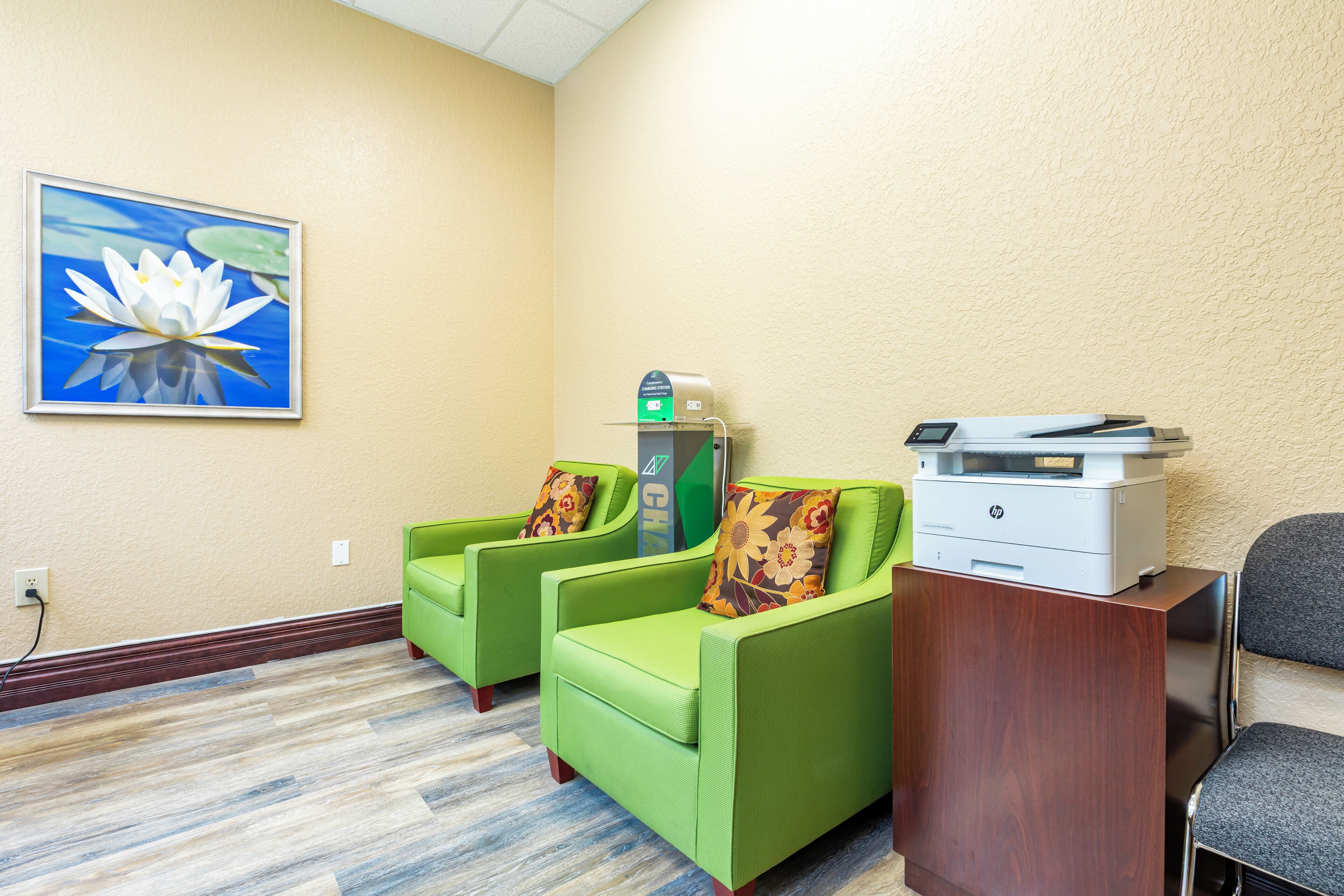 A hotel room with a wooden floor, green armchairs, a desk with a printer, and a wall-mounted trash can.