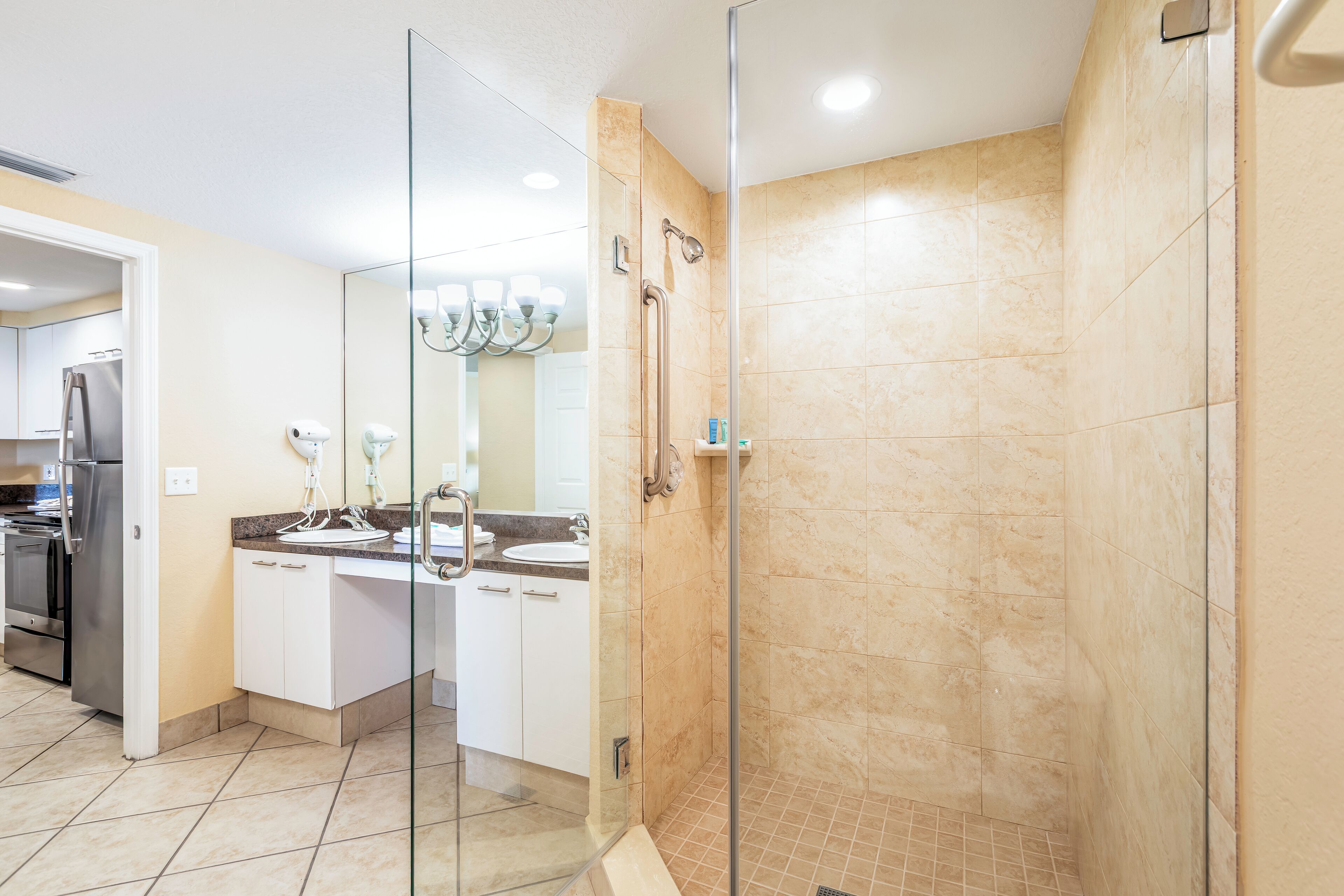 A modern kitchen with white cabinets, a stainless steel refrigerator, and a glass-enclosed shower area with beige tiles.