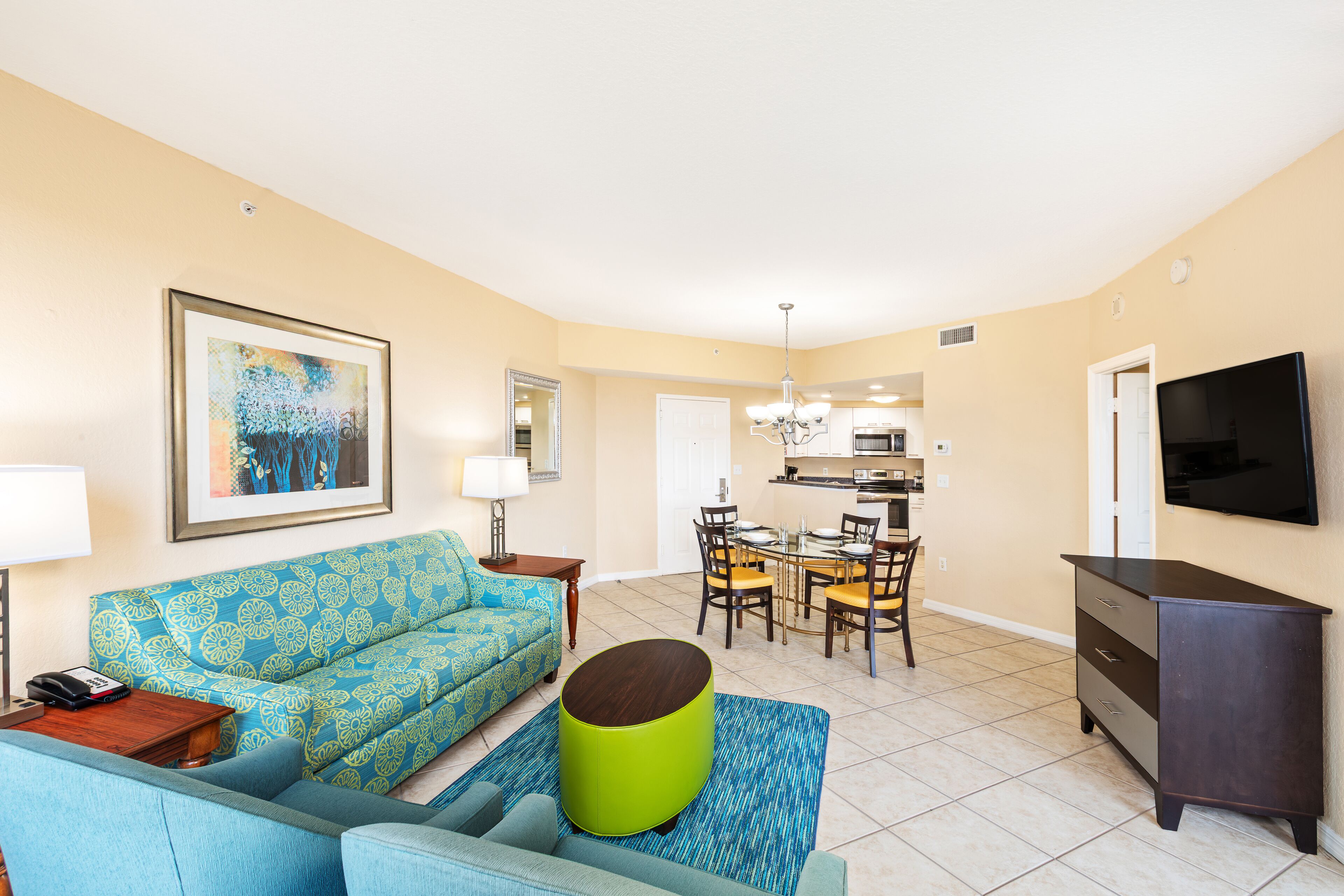 A living room with a sofa, a coffee table, a dining area with chairs and a table, a kitchen area, and a TV mounted on the wall.