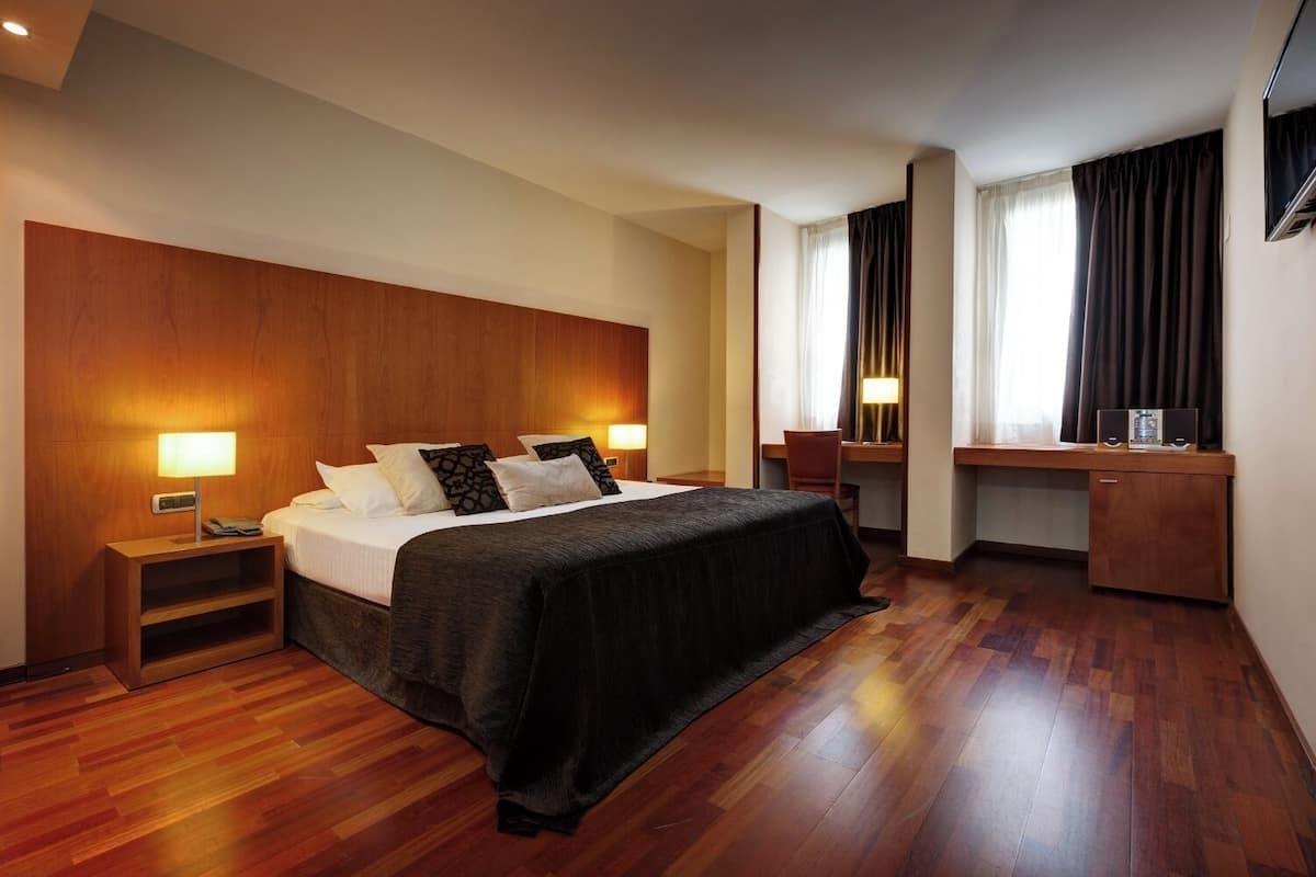 A hotel room with a wooden headboard, a bed with a black bedspread, two bedside tables with lamps, a desk with a chair, and a TV mounted on the wall.