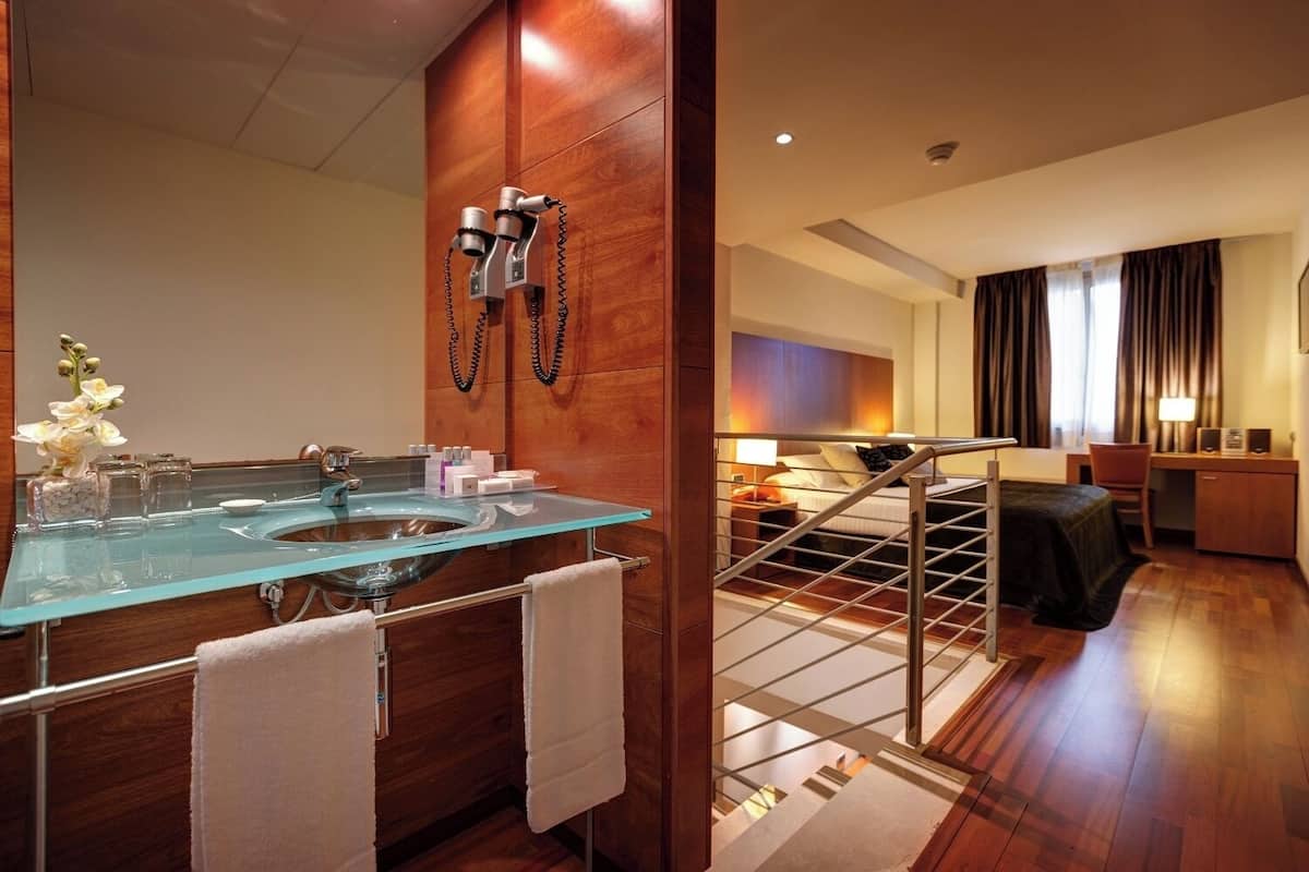 A modern bathroom with a glass sink, wooden vanity, and a mirror. There is a staircase leading to a bedroom with a bed, desk, and a view of the outside.
