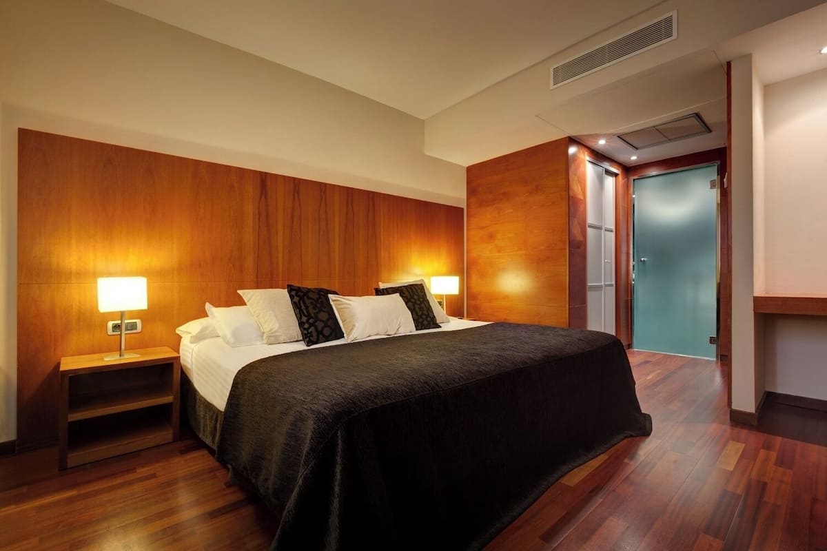 A hotel room with a large bed, wooden panel walls, two bedside lamps, and a door leading to another room.