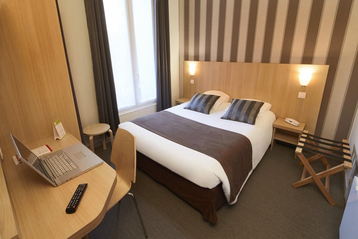 A hotel room with a bed, a desk with a laptop, a chair, a bedside table with a lamp, and a window with curtains.