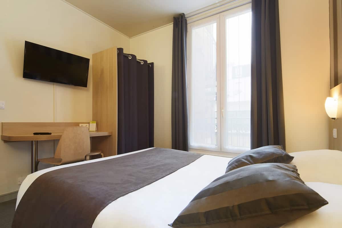 A hotel room with a bed, a desk, a chair, a TV mounted on the wall, and a window with curtains.