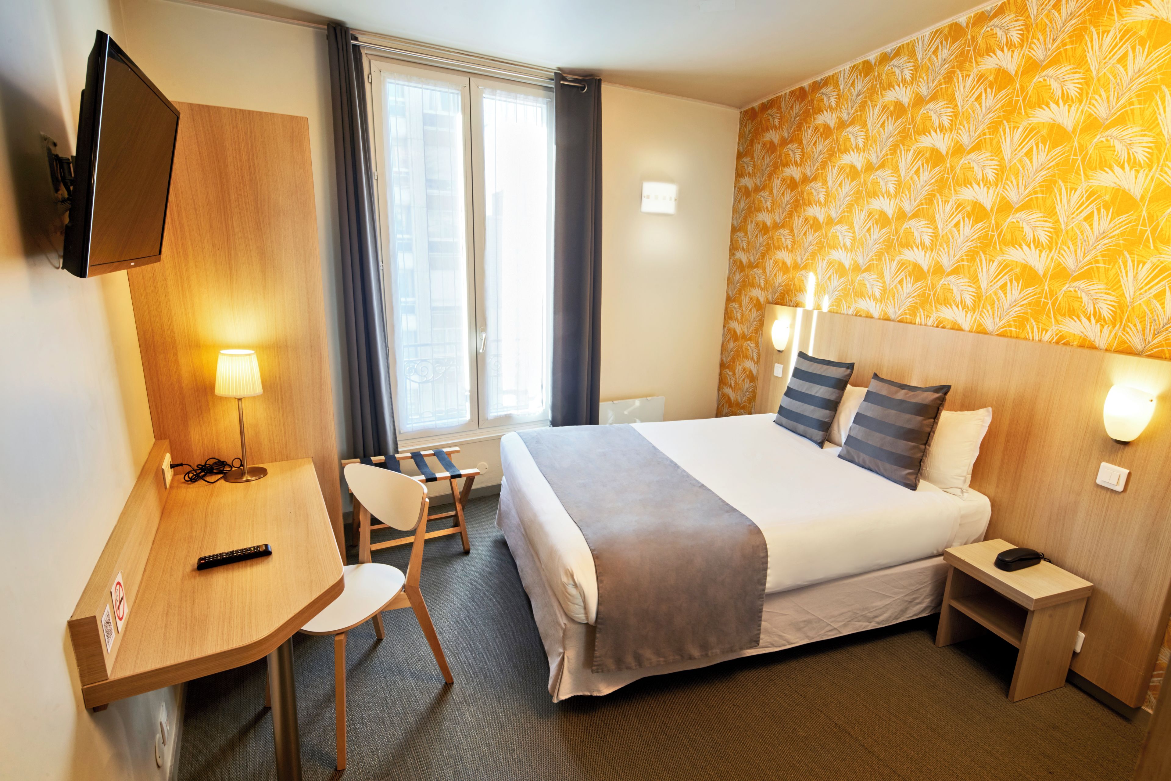 Habitación de hotel con cama, mesitas de noche, escritorio con silla, televisor montado en la pared y ventana con cortinas.