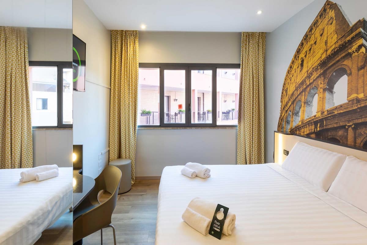 A hotel room with two beds, a desk, a chair, and a large window with curtains.