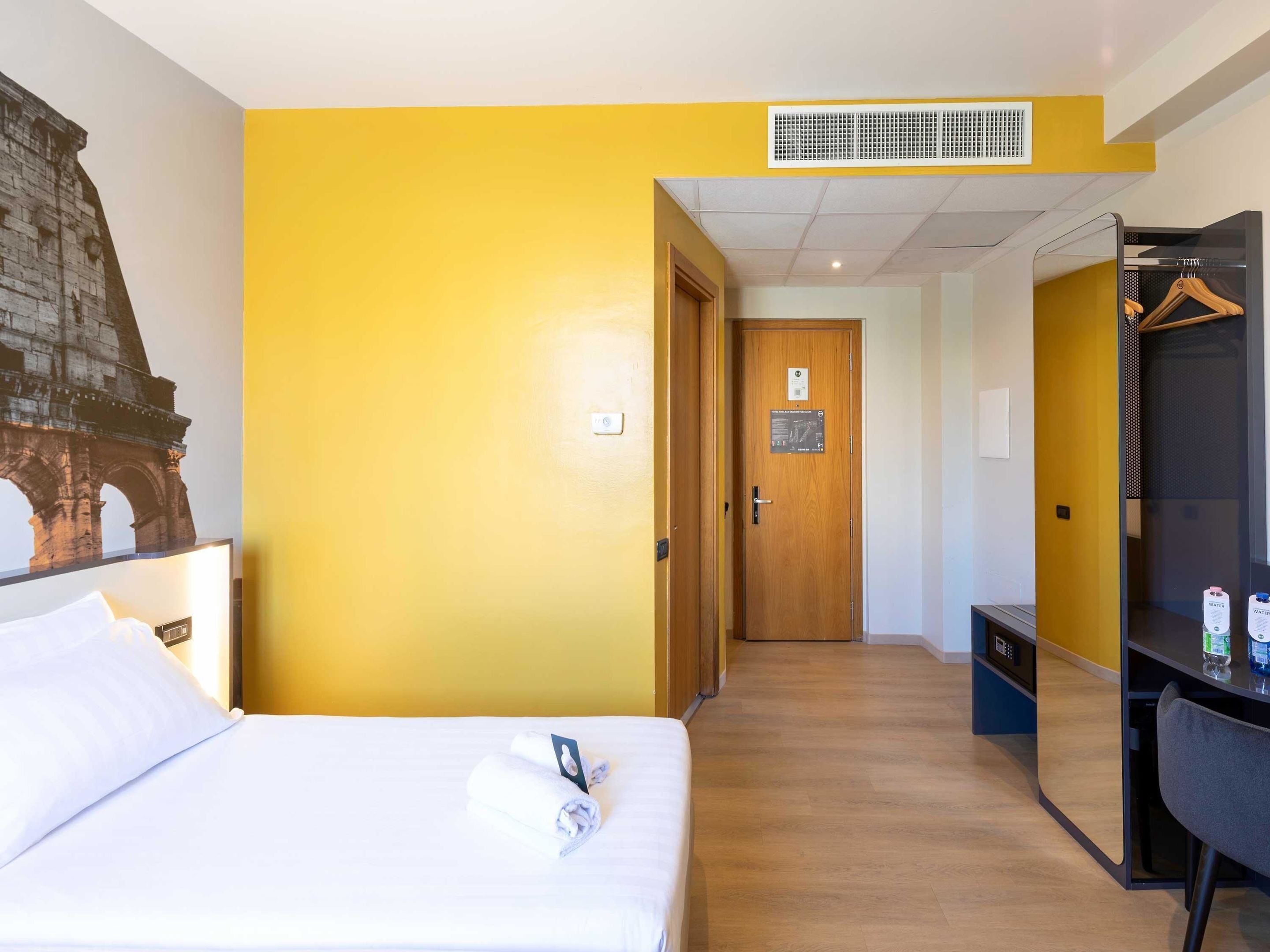 A hotel room with a bed, a wooden door, a wardrobe, and a wall-mounted heater.