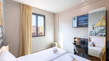 Ein Hotelzimmer mit zwei Betten, einem Schreibtisch, einem Sessel, einem an der Wand montierten Fernseher und einem Fenster mit Vorhängen.