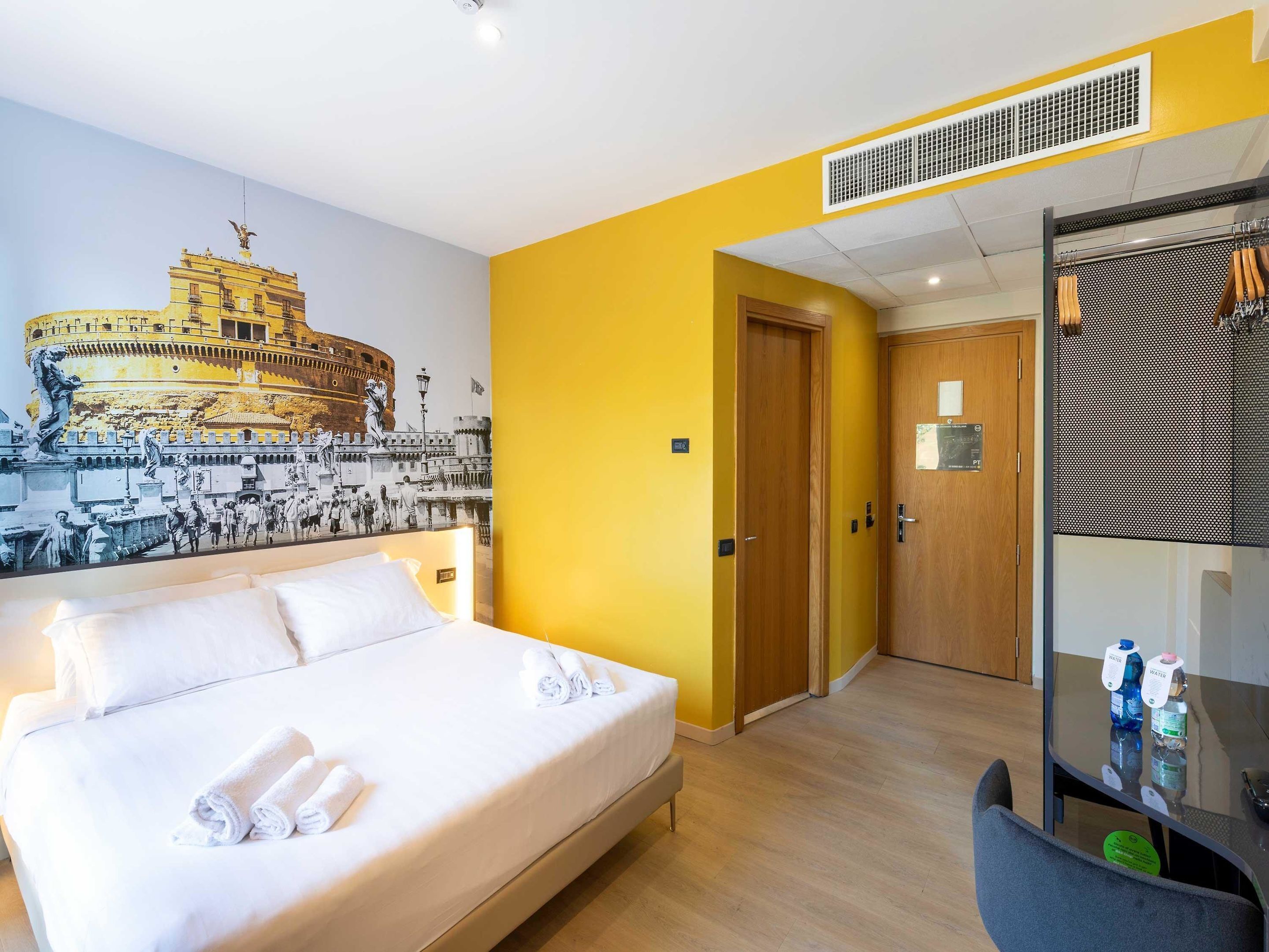 Habitación de hotel con una cama, un mural de un edificio histórico, una puerta de madera y un espejo con una botella de agua sobre la mesa.