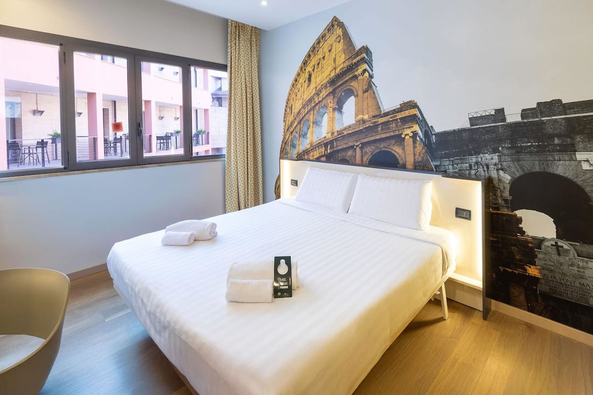 A hotel room with a large bed, a headboard with a mural, a window with a view of the outside, and a chair.