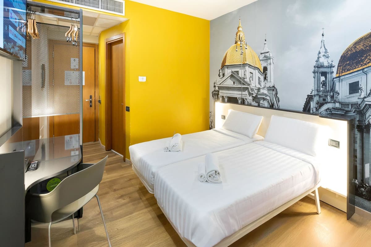 A hotel room with a bed, a desk, and a chair. There is a painting of a building on the wall.