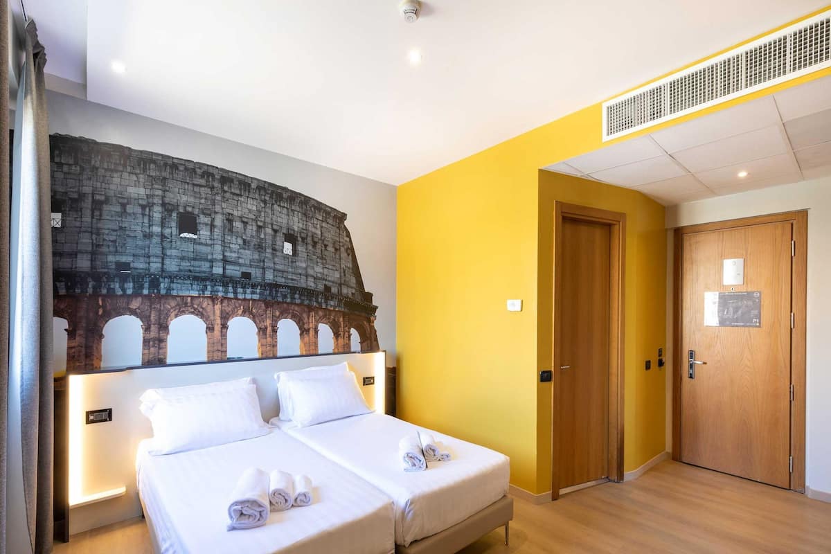 A hotel room with two beds, a large wall mural of the Colosseum, a wooden door, and a ceiling vent.