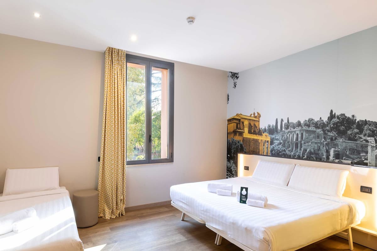 A hotel room with a bed, a sofa, a window with curtains, and a mural of an ancient city.