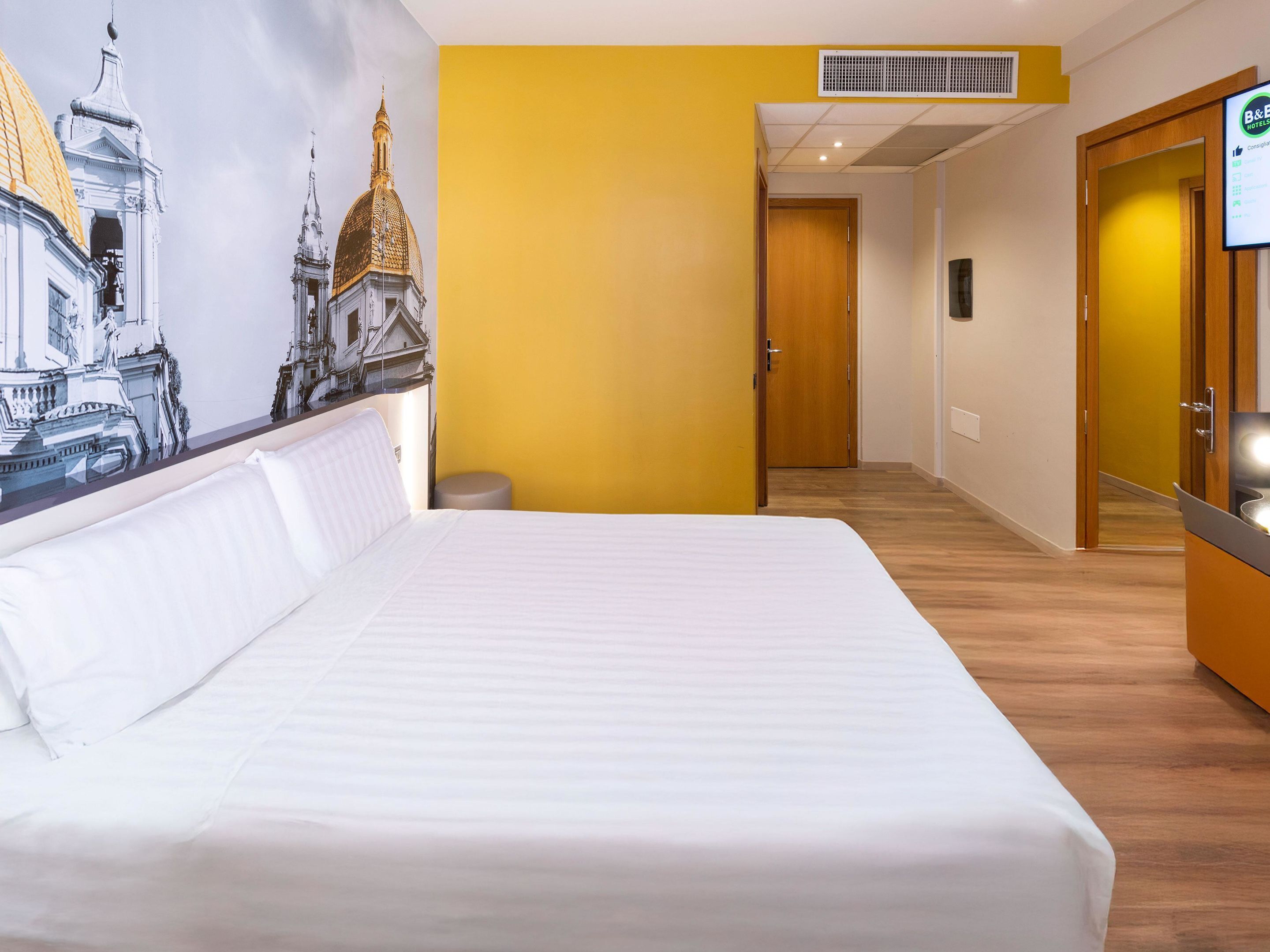 Habitación de hotel con una cama grande, un mural de un edificio, piso de madera y una puerta que da a otra habitación.