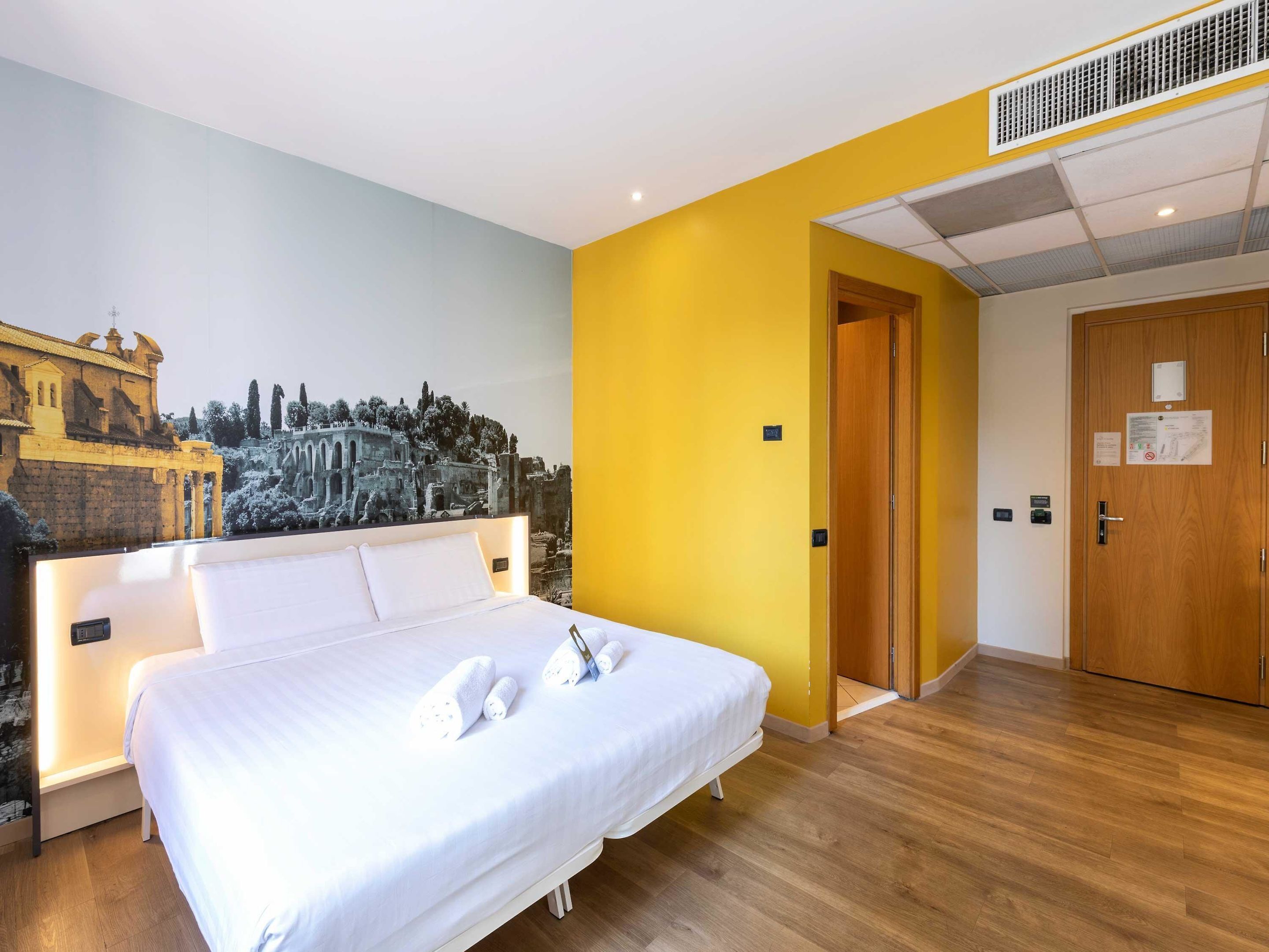 Una habitación de hotel con una cama grande, un mural de ruinas antiguas, una puerta de madera y una pared de acento amarilla.