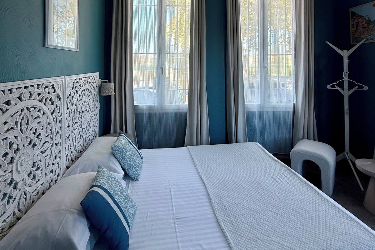 A bedroom with a large bed, two windows with curtains, a white headboard, and a small stool.