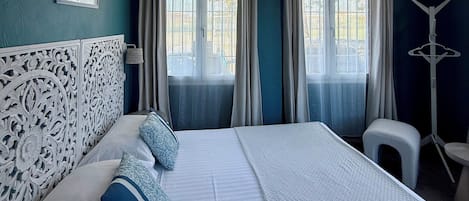 Un dormitorio con una cama grande, dos ventanas con cortinas, un cabecero blanco y un pequeño taburete.